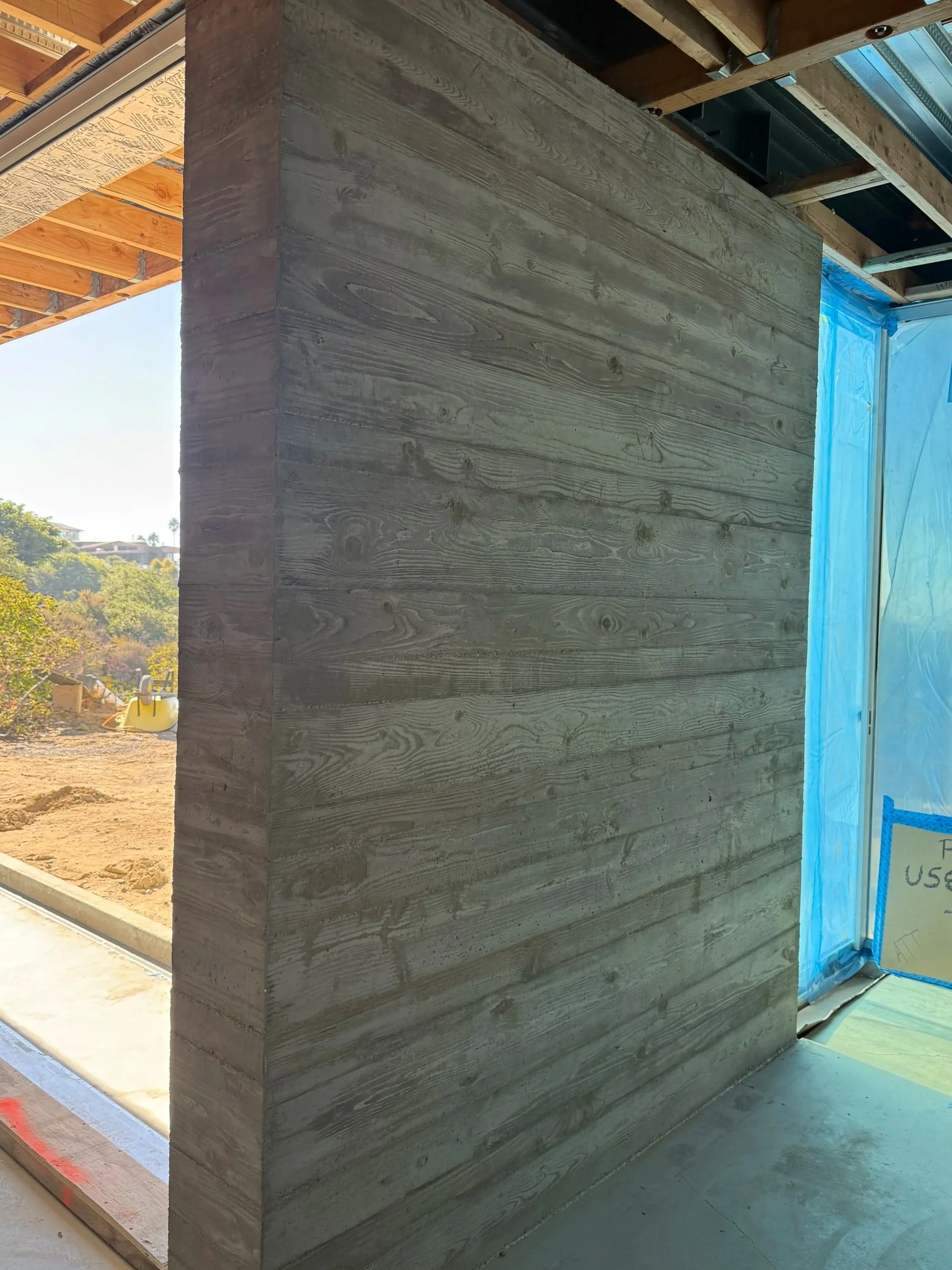 Interior view of an under-construction building featuring a partially built wooden wall with visible horizontal wood planks. Outside scenery shows trees and construction equipment.