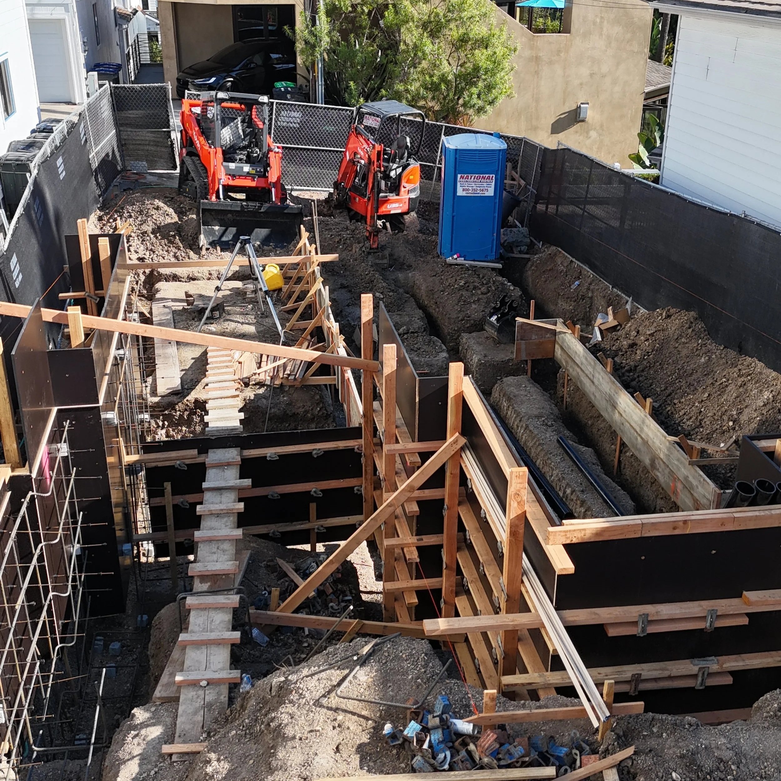 Construction site with deep excavations and wooden formwork for concrete, two small excavators, a portable toilet, and nearby residential buildings.