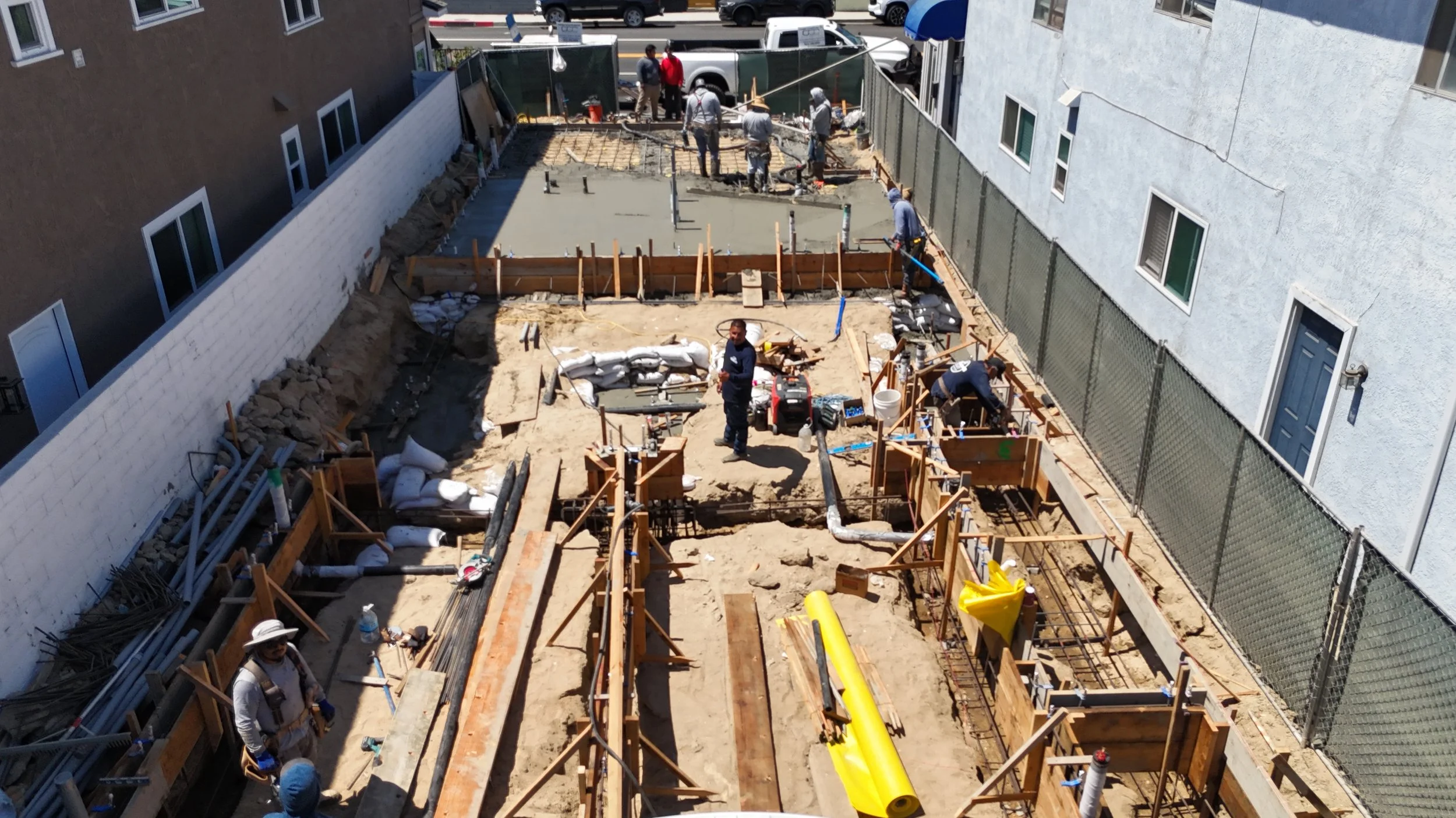 Construction workers working on a building foundation in a narrow lot surrounded by residential buildings.