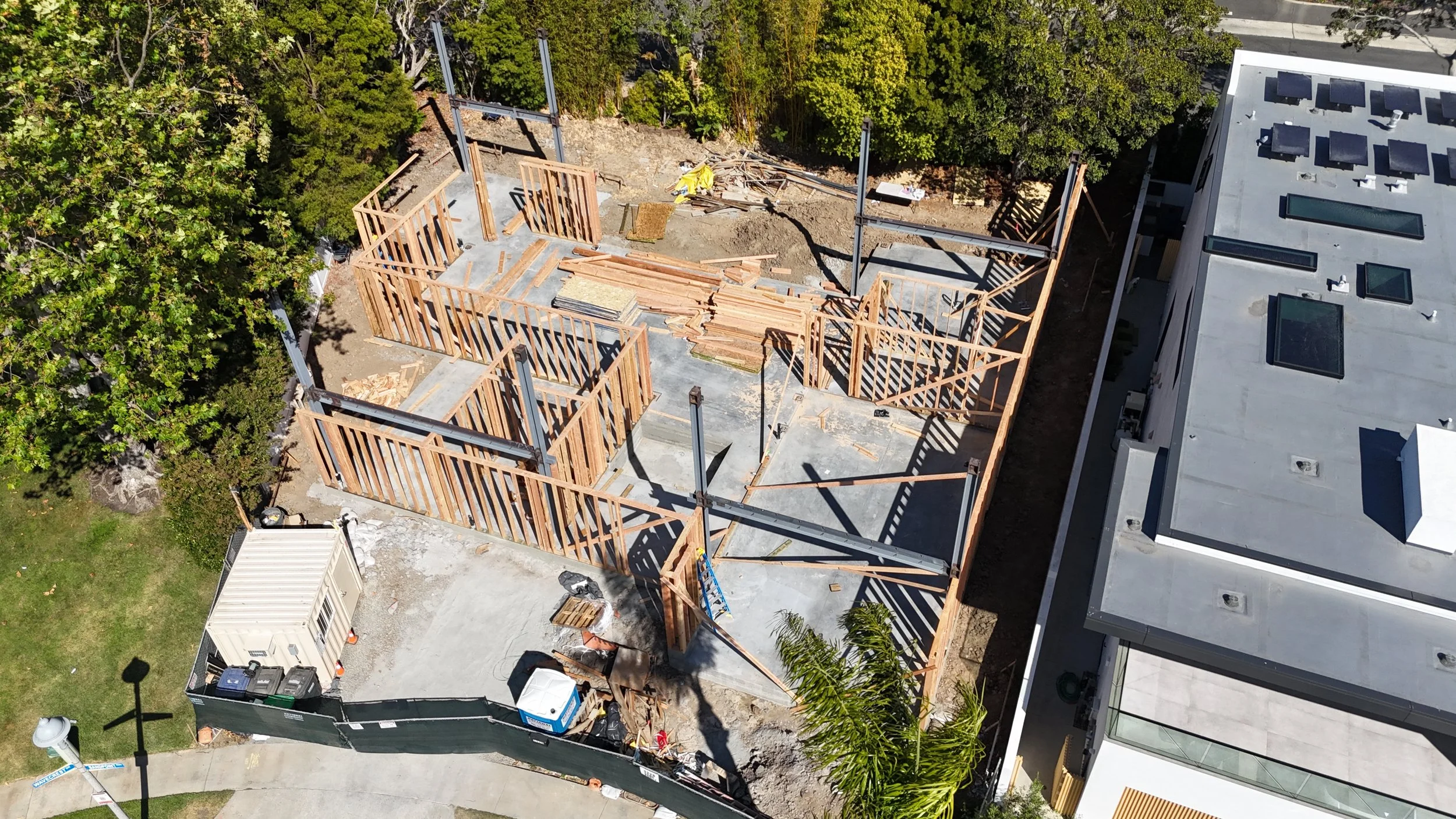 Aerial view of a construction site with wooden framing for a building, nearby trees, and an adjacent modern flat-roofed building.