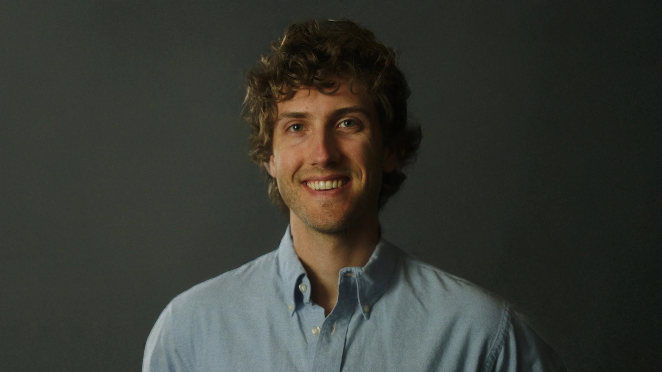 Man with curly hair and blue button down shirt smiling.