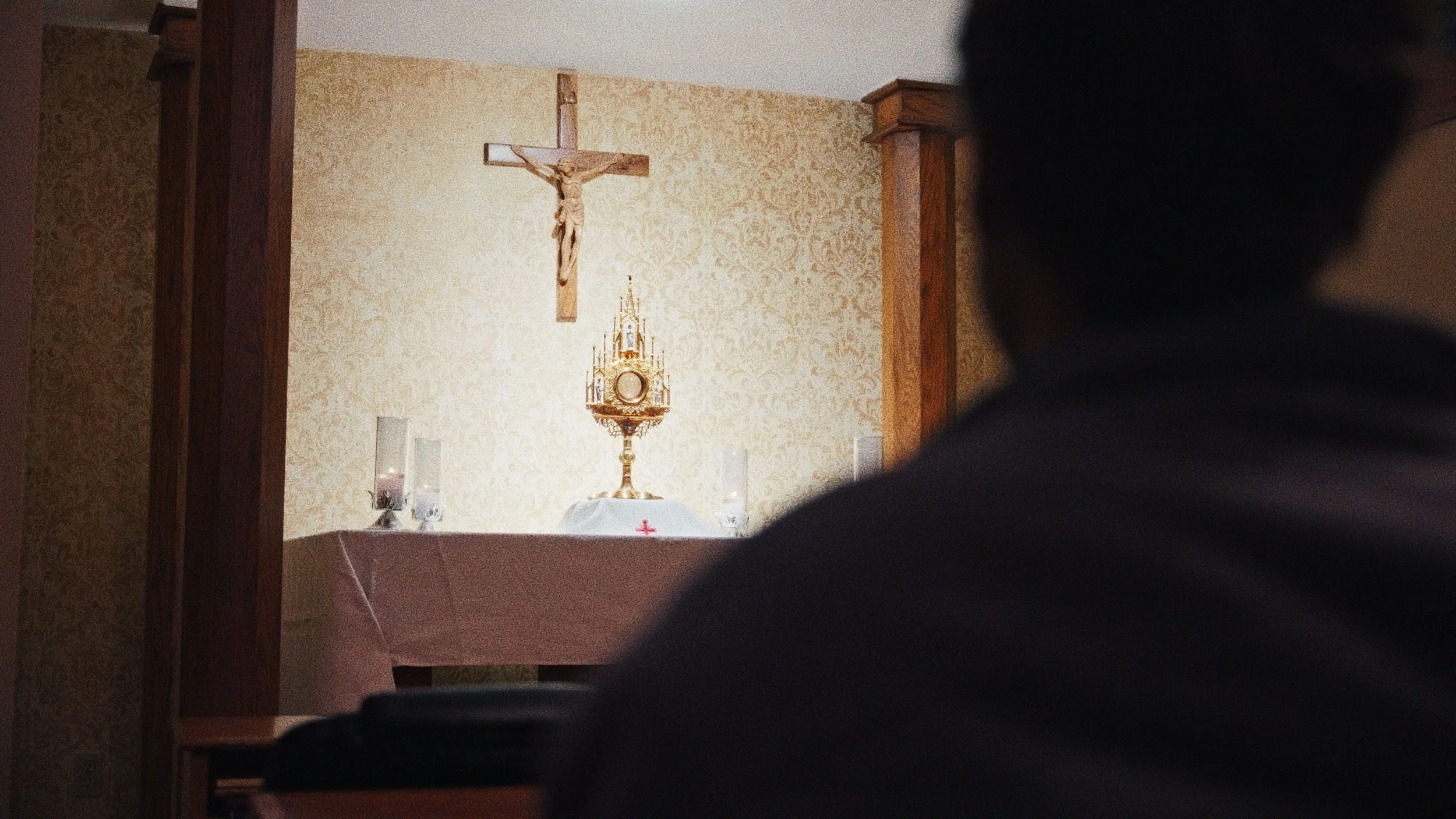Inside a Catholic chapel, a crucifix on the wall and the Eucharist in the monstrance on the altar, with a person sitting nearby facing the altar.