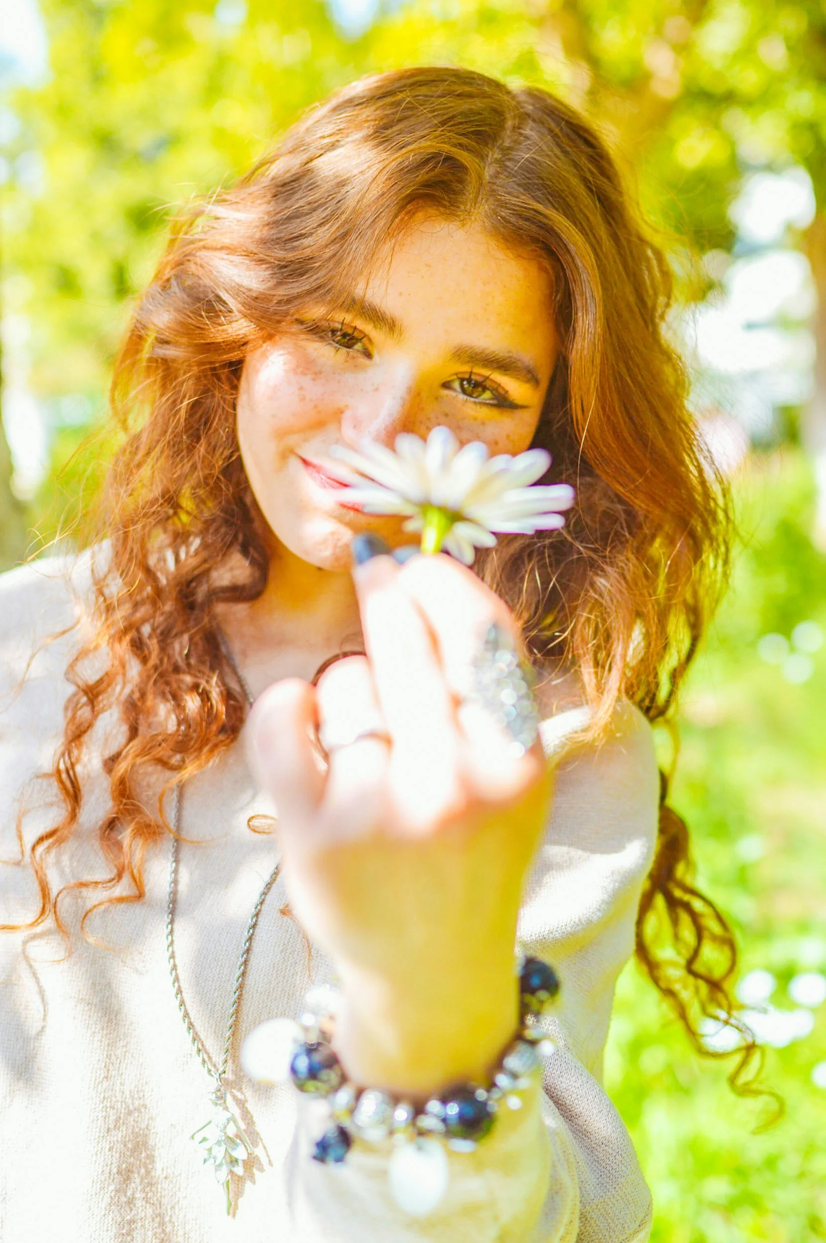 Young woman with curly red hair and freckles holding a white daisy flower, outdoors in a sunlit park with green trees in the background.