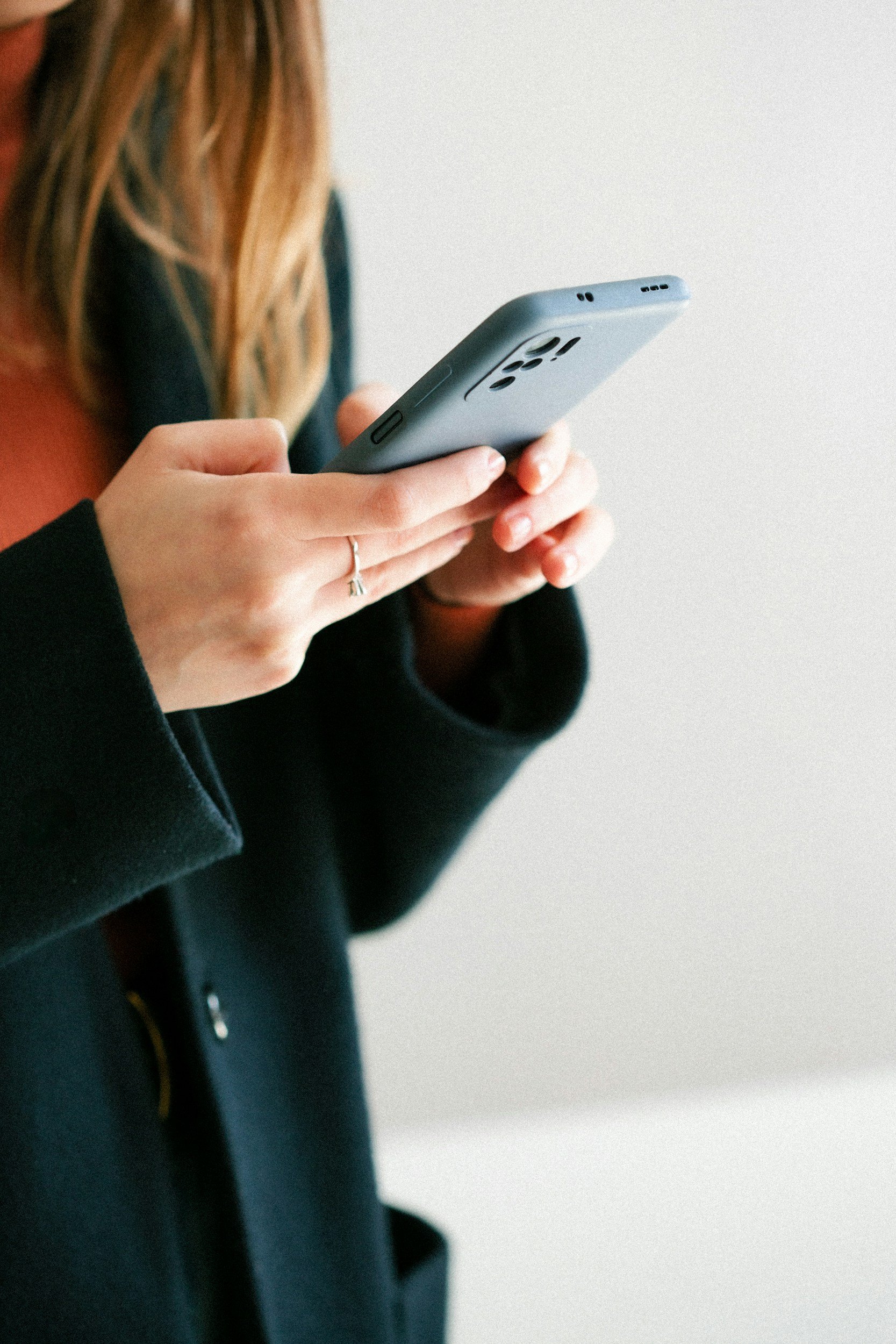 Close-up of a person with long hair holding a smartphone, wearing a black jacket, against a plain, light background.