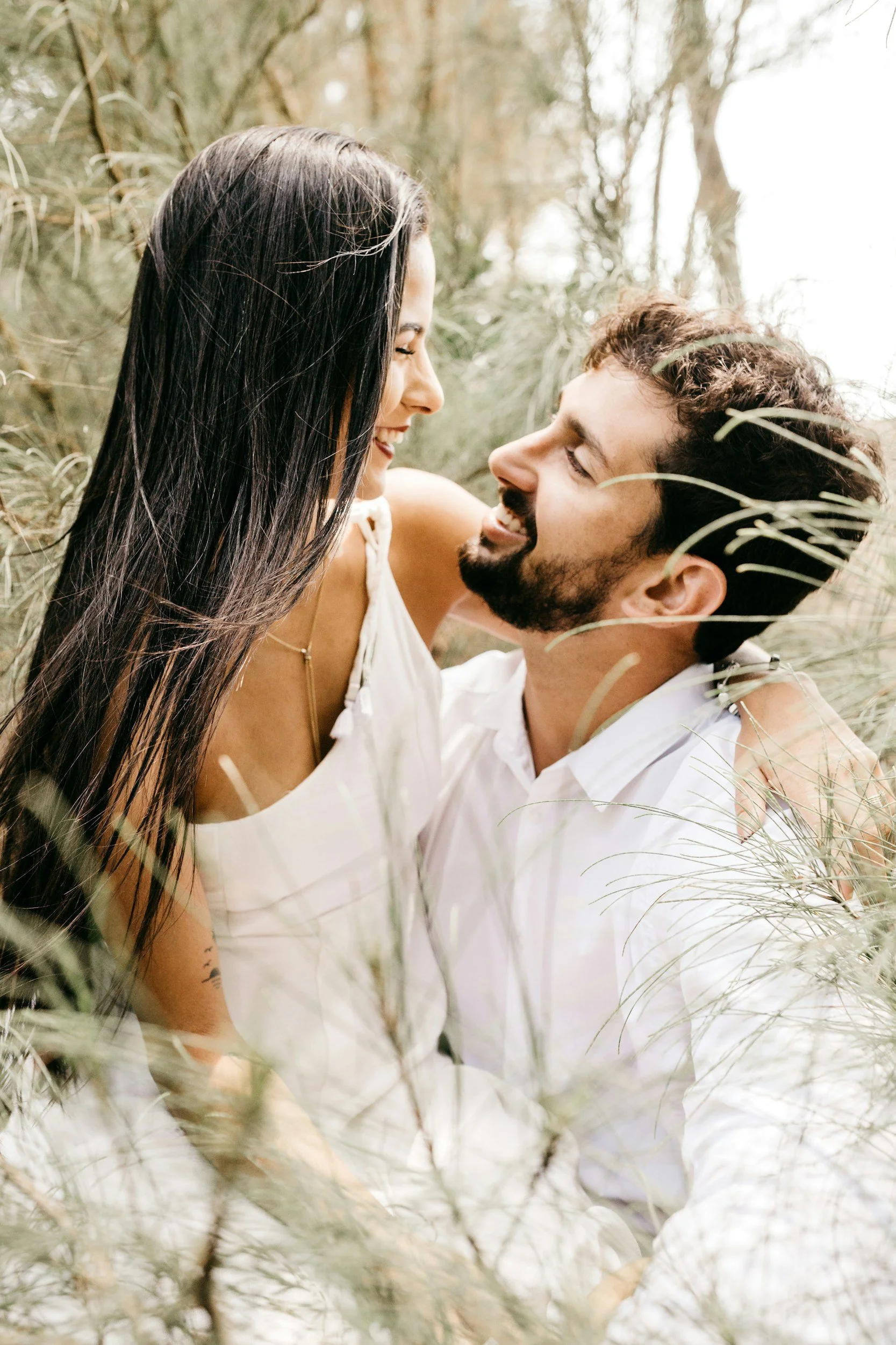 A couple smiling and looking into each other's eyes while lying in a grassy field during daylight.