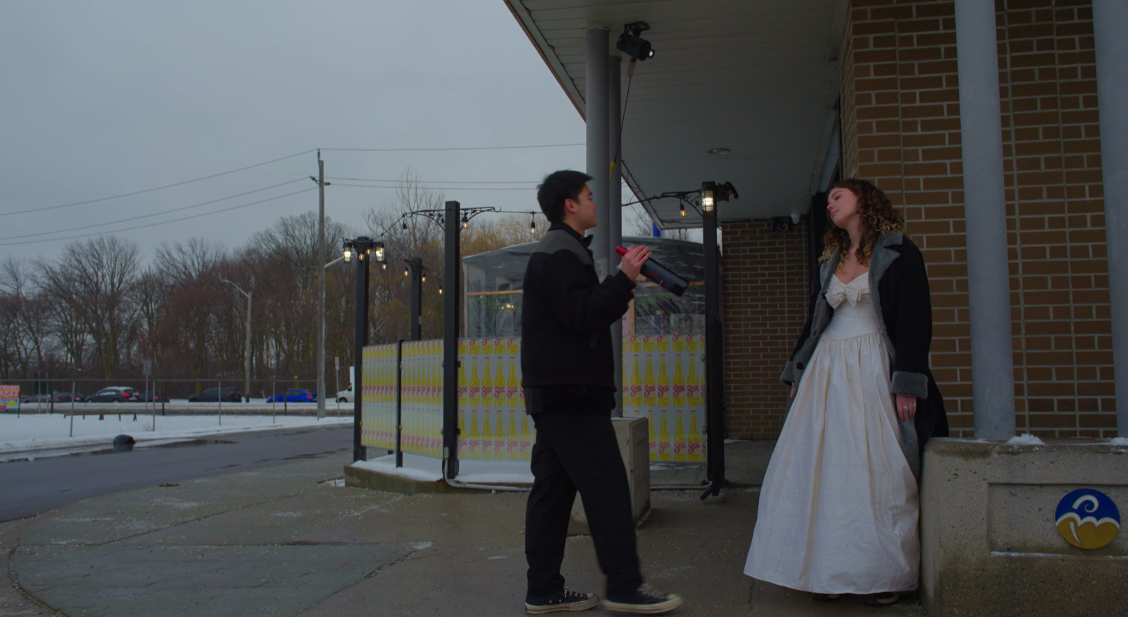 A young man holding a microphone talking to a young woman in a white dress and black coat outside a building with brick walls and a sidewalk, overcast weather.
