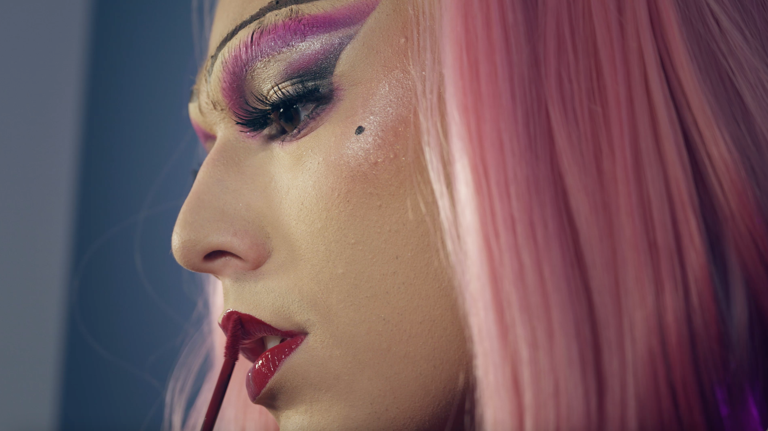 A close-up of a woman with pink hair applying red lipstick.