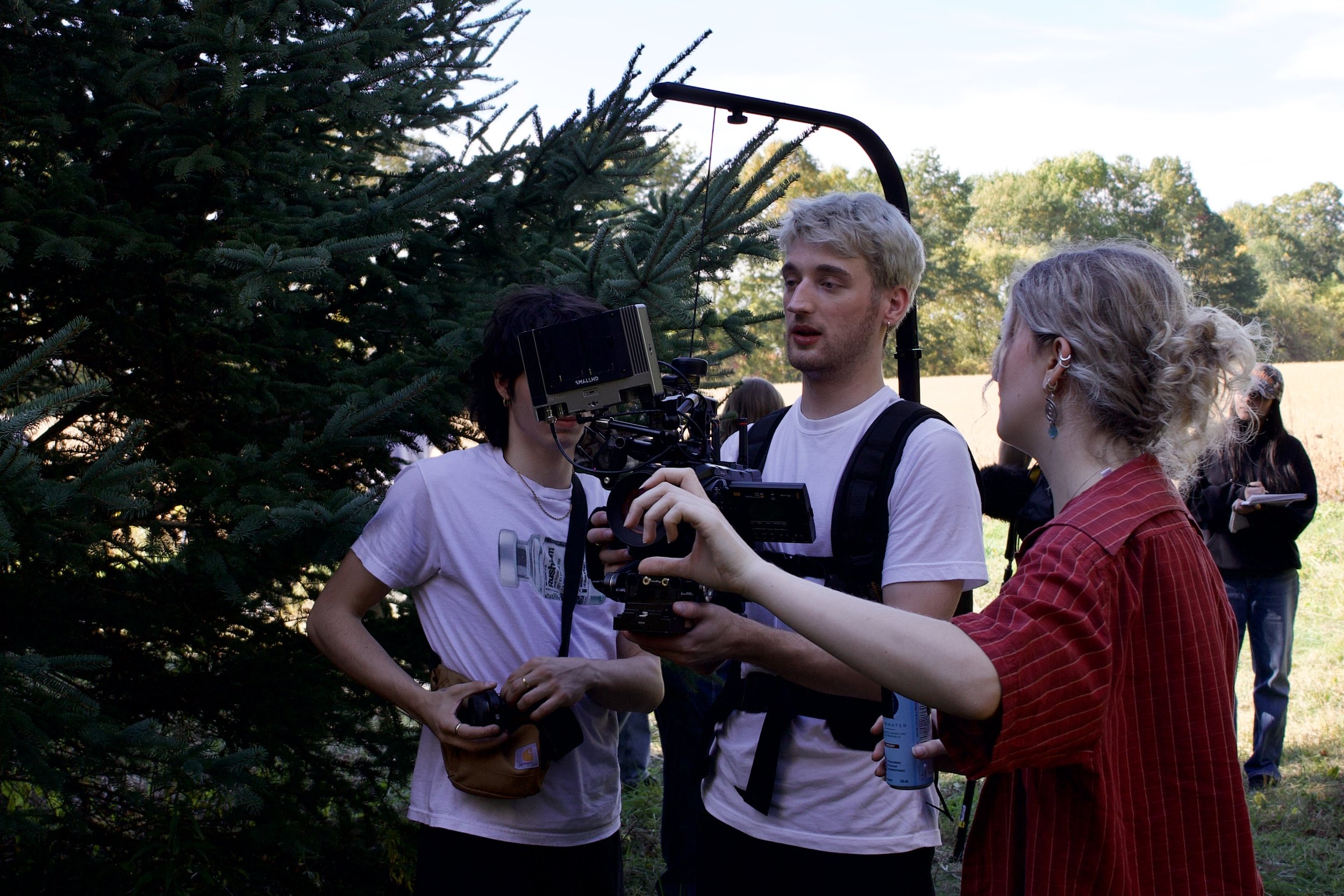 Three people filming outdoors near a large evergreen tree during daylight, with a woman in a red shirt pointing a camera at a young man with blonde hair, while another young man stands nearby.