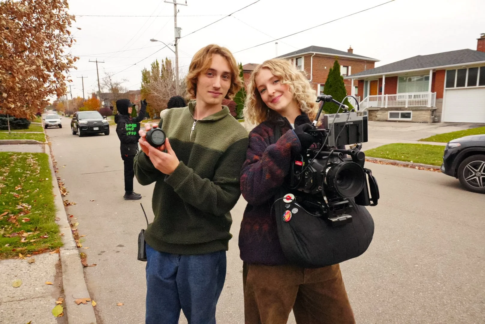 Two young people, a male and a female, stand in the street with film equipment. The male holds a camera lens, and the female operates a professional film camera. There are other people and cars in the background, and houses line the street with autumn trees.