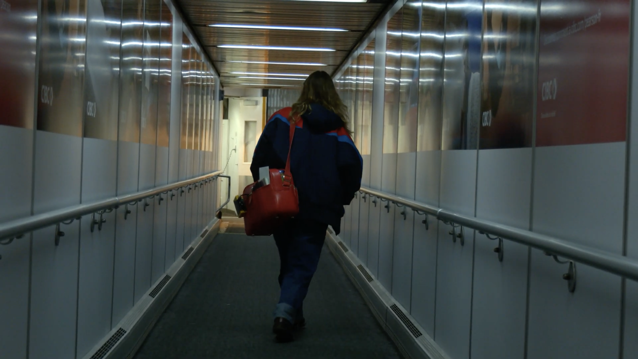 A woman walking through an airport jet bridge carrying a red bag, wearing a dark jacket and jeans, with lighting above.