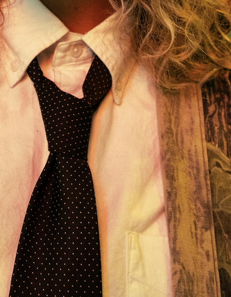 Close-up of a person wearing a light-colored blazer, a white collared shirt, and a black tie with small white polka dots, with curly blond hair visible.