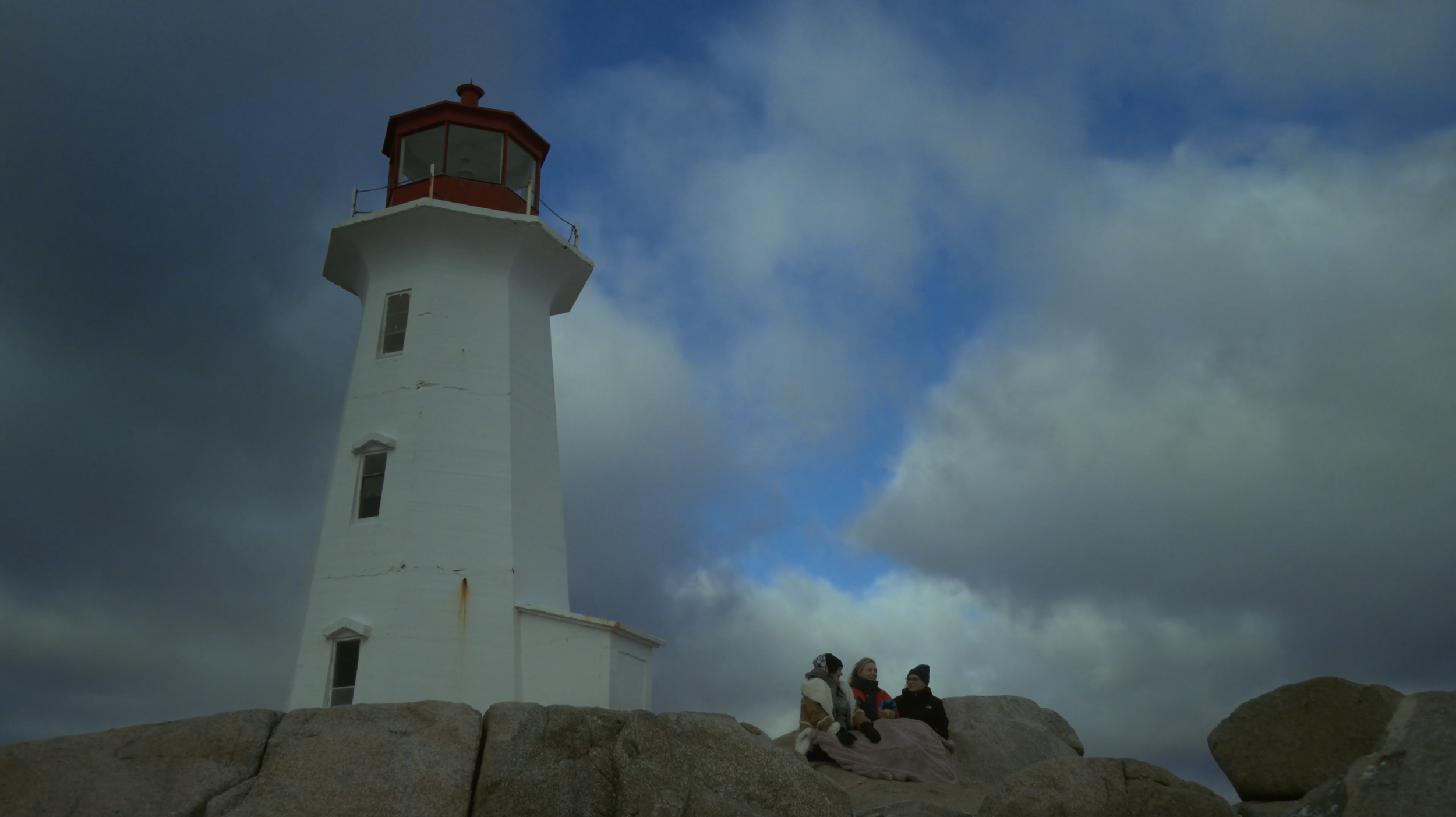 A white lighthouse with a red top stands on a rocky shore under a cloudy sky, with three people sitting on the rocks nearby.