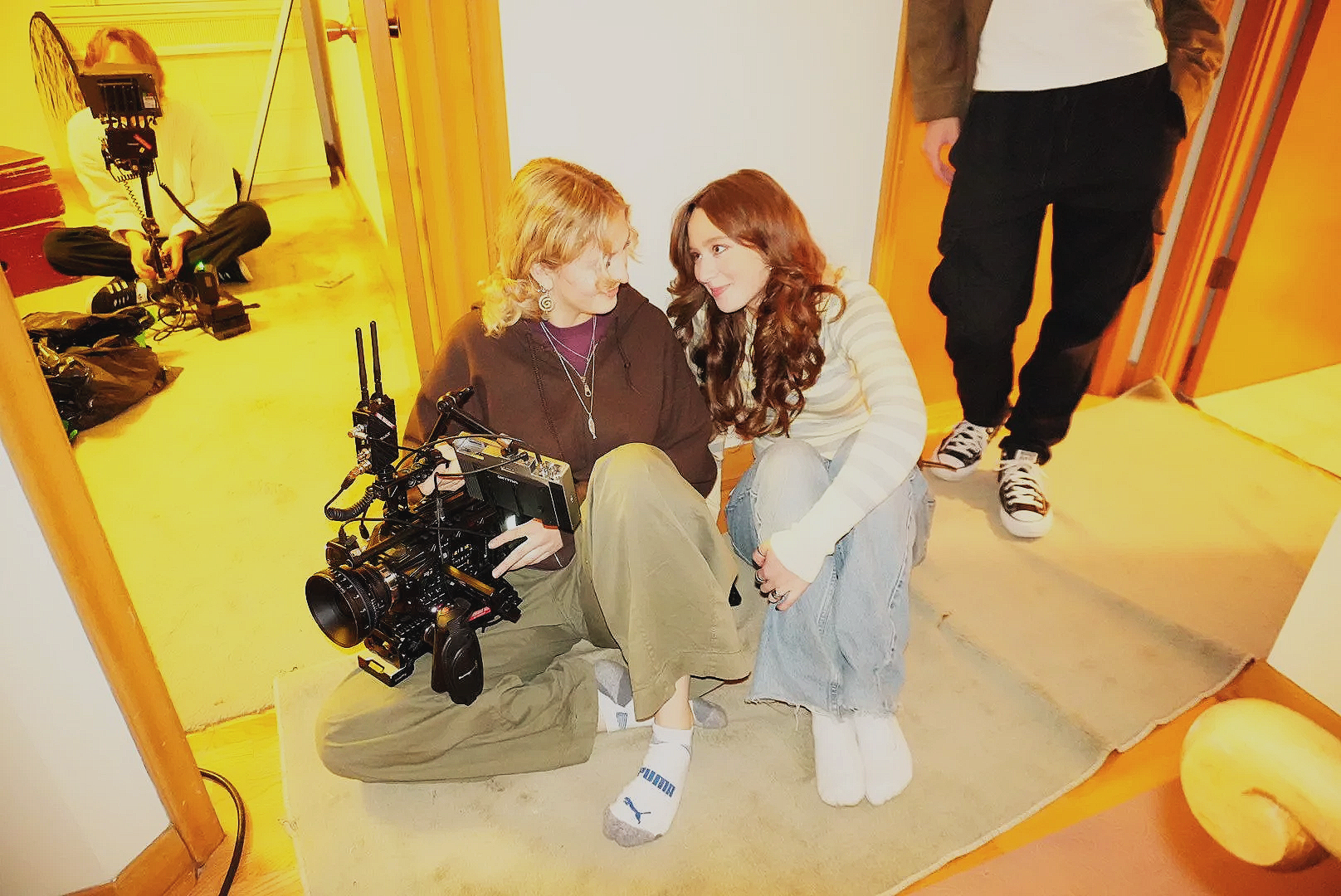 Two women sitting on the floor and engaging in a conversation, with a cameraman sitting behind them in a room with wooden walls. A man is standing nearby, partially out of the frame.