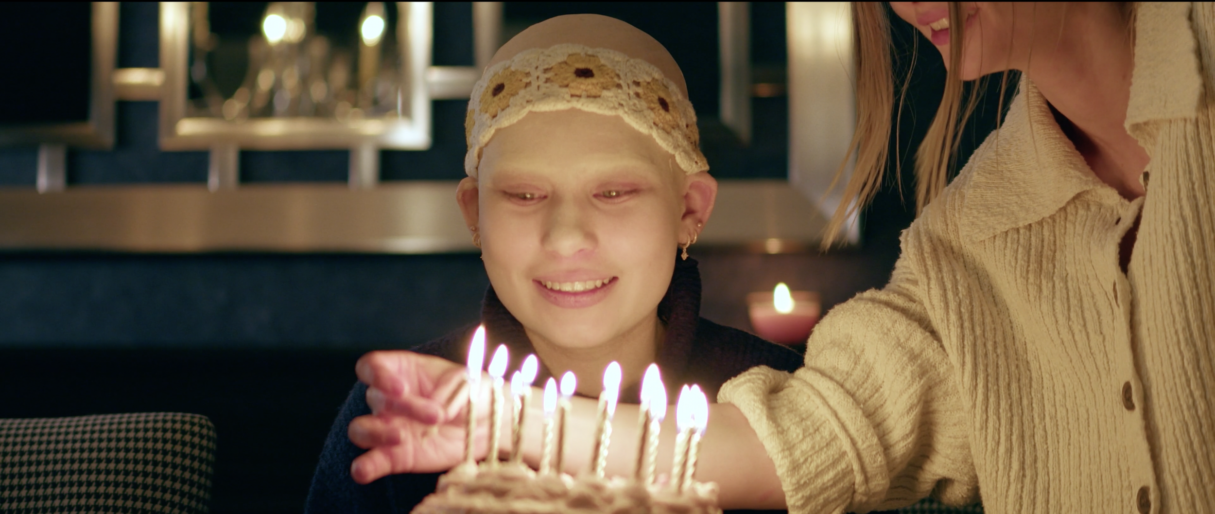 A young girl with a bandage on her head, smiling, as a woman helps her blow out candles on a birthday cake.