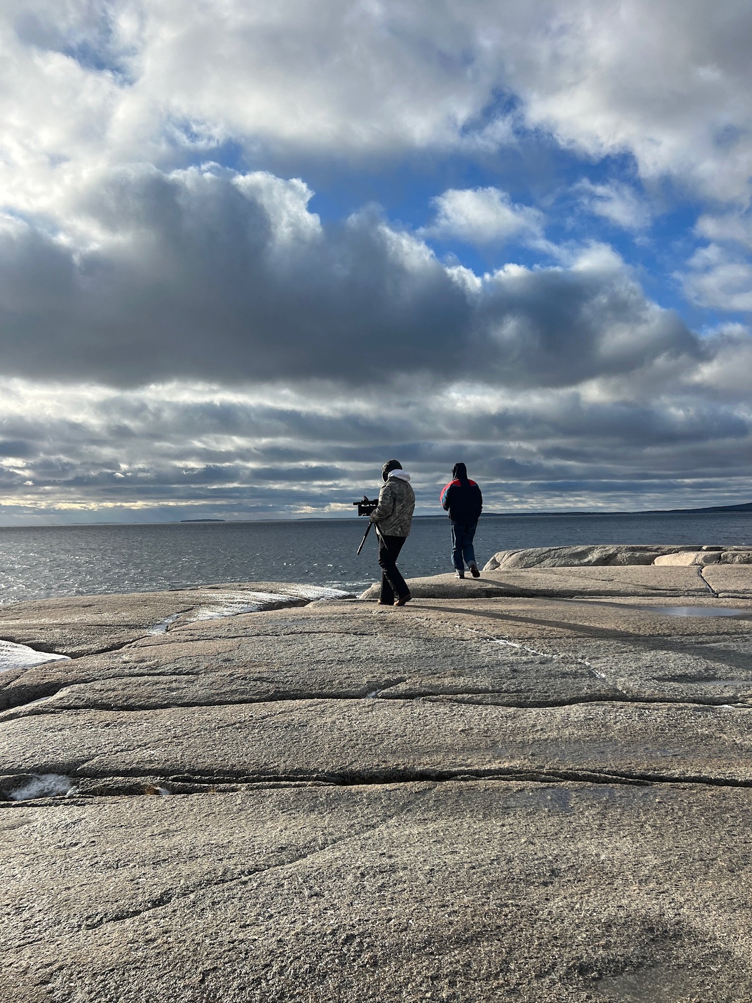 Two people walk along a rocky shoreline beside a large body of water under a cloudy sky.