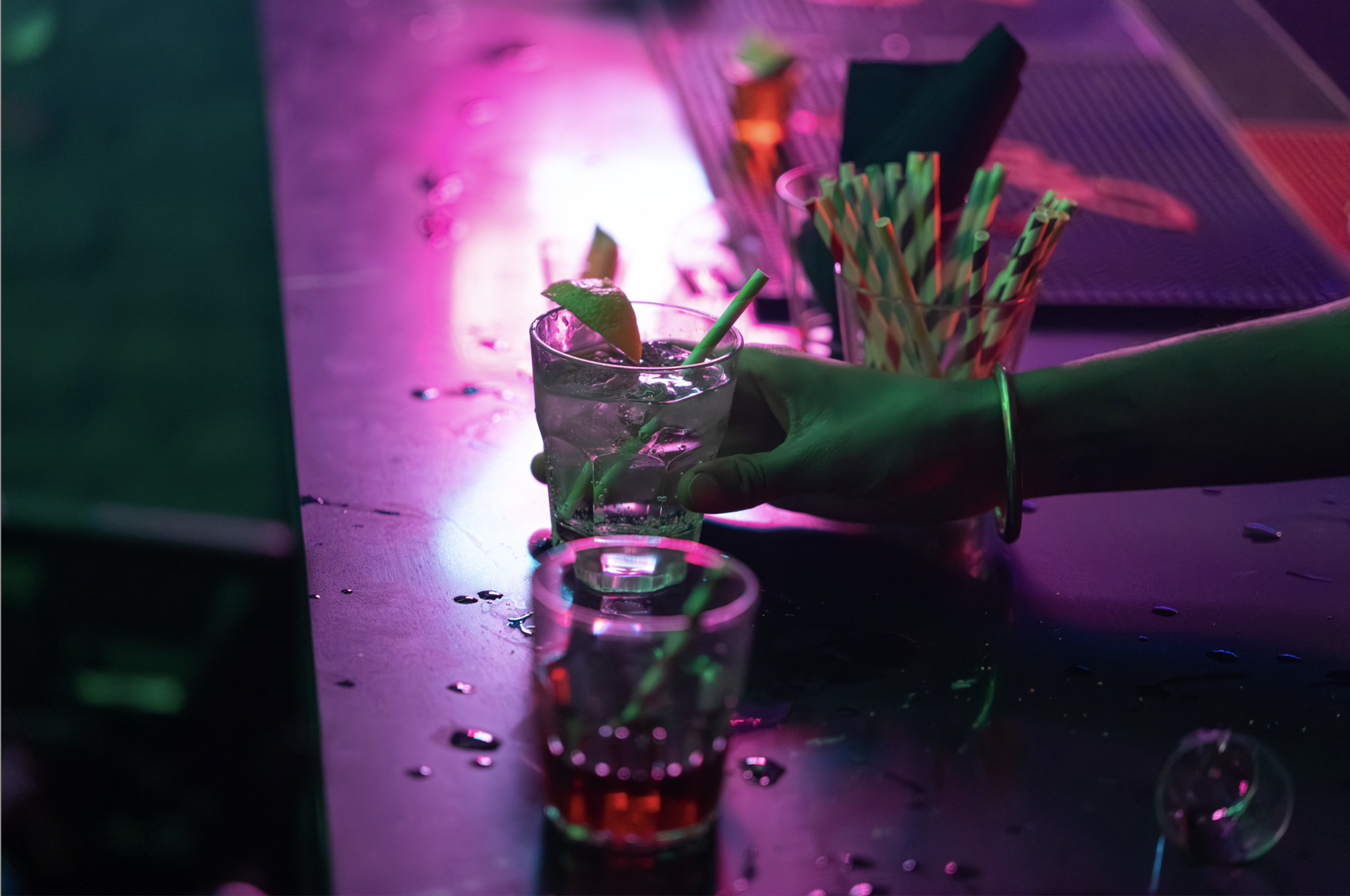 Person holding a shot glass with a lime wedge, surrounded by more shot glasses, colorful straws, and a neon pink and purple glowing bar scene.