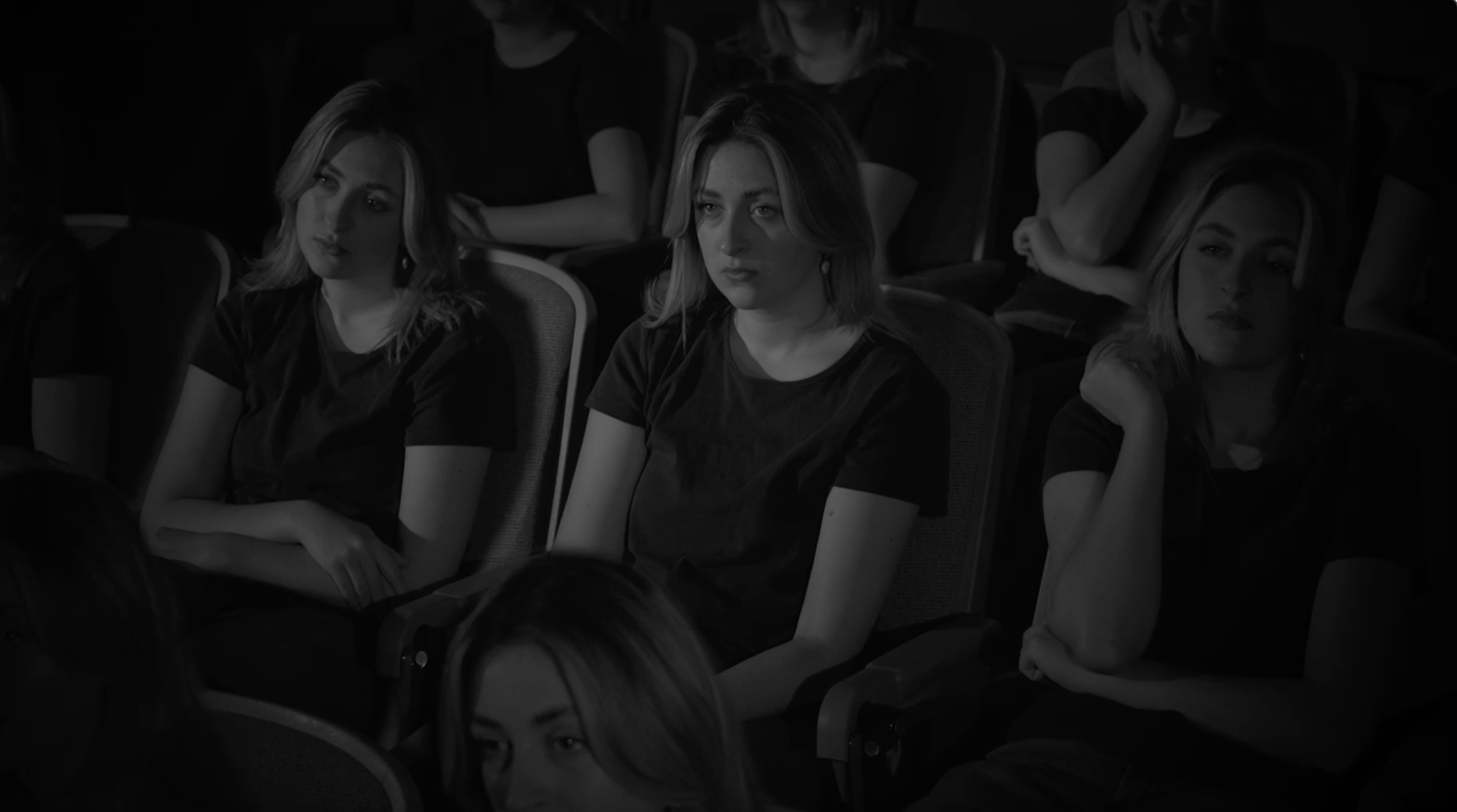 A group of young women sitting in an auditorium, appearing attentive, with one woman in the foreground and others behind her, all wearing dark clothing.