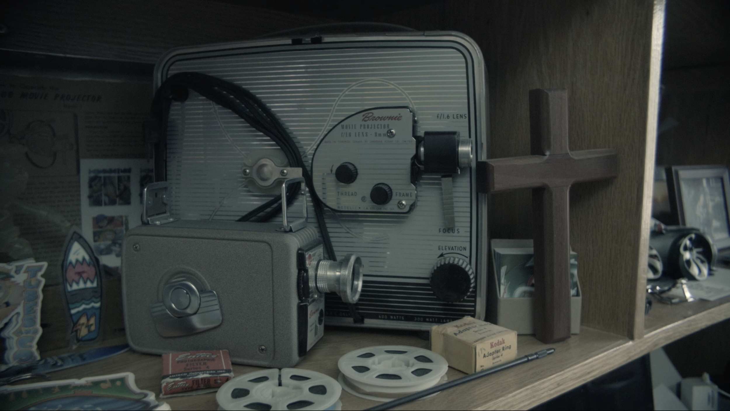 A vintage movie projector on a wooden shelf surrounded by memorabilia, cross-shaped decoration, film reels, and miscellaneous items.