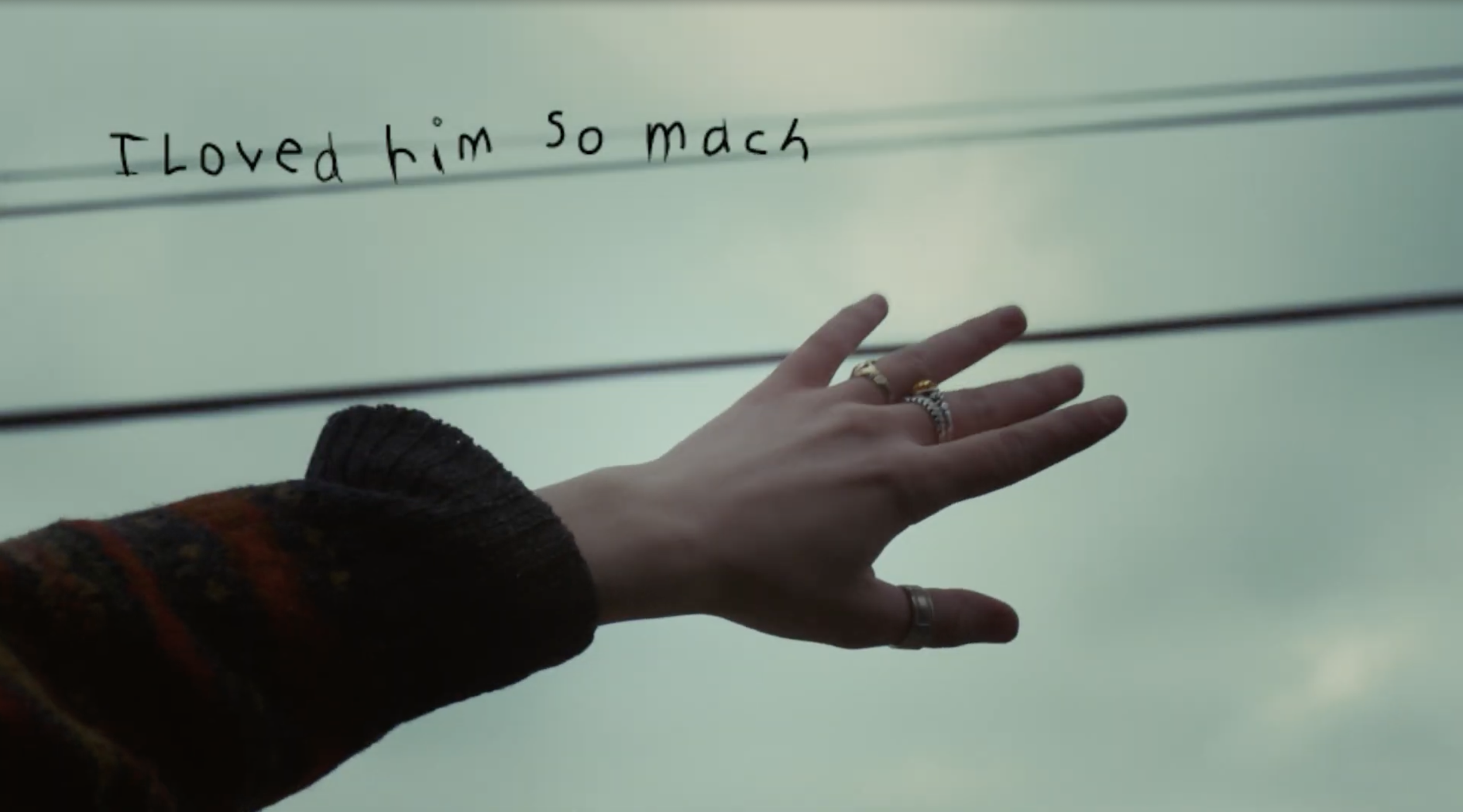 Close-up of a hand with rings reaching towards a note that says 'I loved him so much' written on a white surface against a pale sky background.