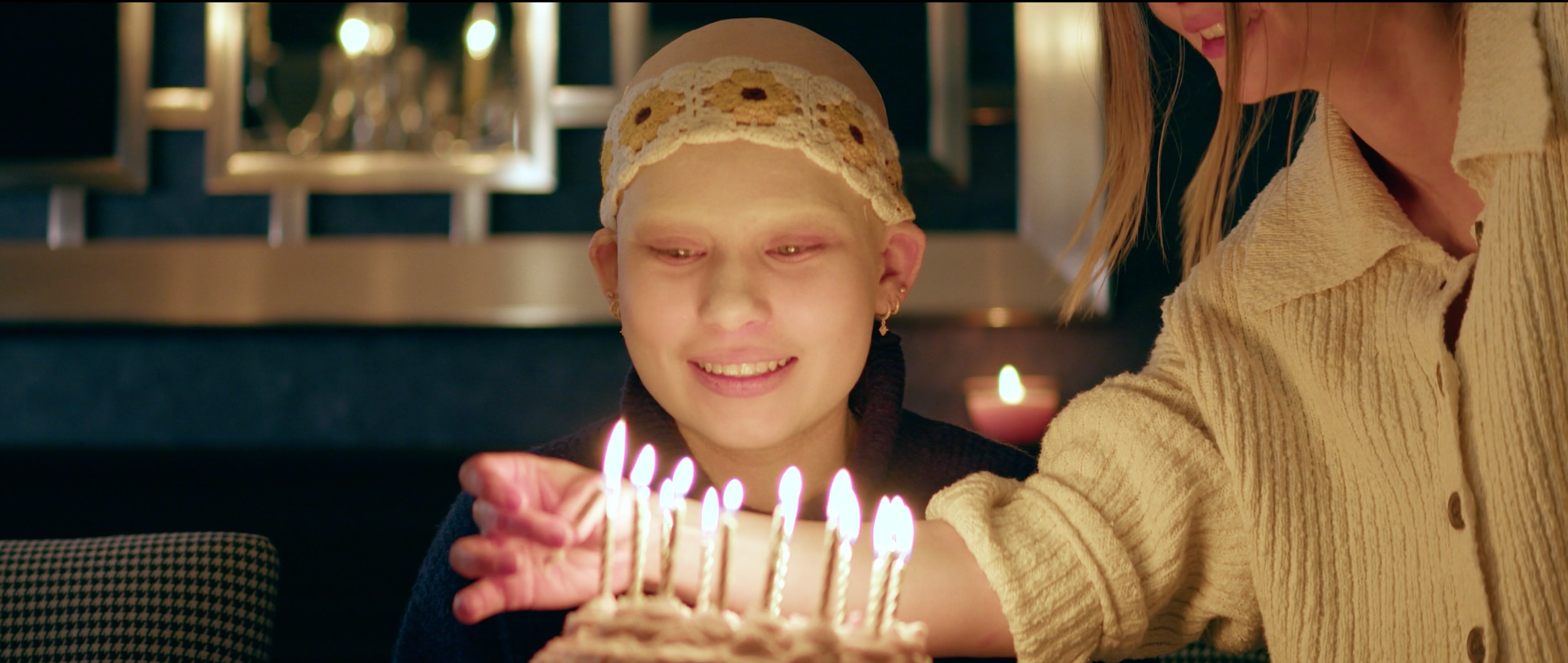 A girl with a bandage on her head smiling at a birthday cake with lit candles, accompanied by an adult woman leaning towards her in a cozy setting.