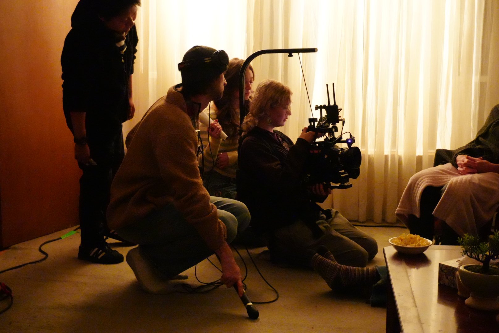 Filmmakers filming a person sitting on a couch in a dimly lit room with curtains, a bowl of snacks, and plants on a table.