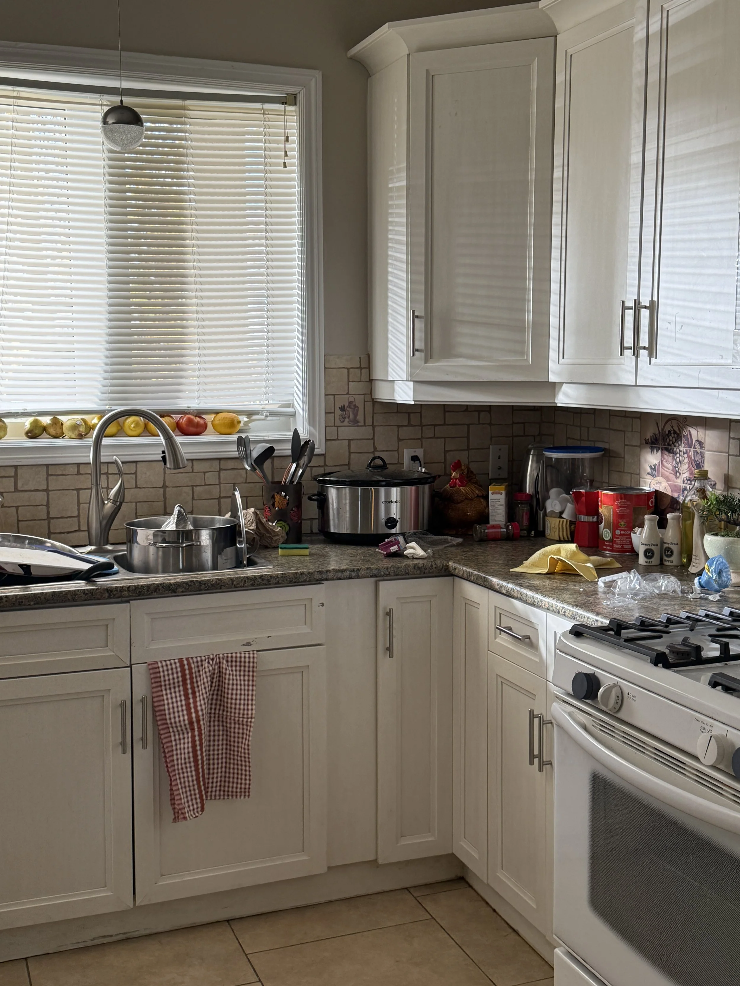 A cluttered kitchen with white cabinets, a granite countertop, and a window with blinds. Various items, including fruit, kitchen utensils, small appliances, and containers, are scattered across the counter.