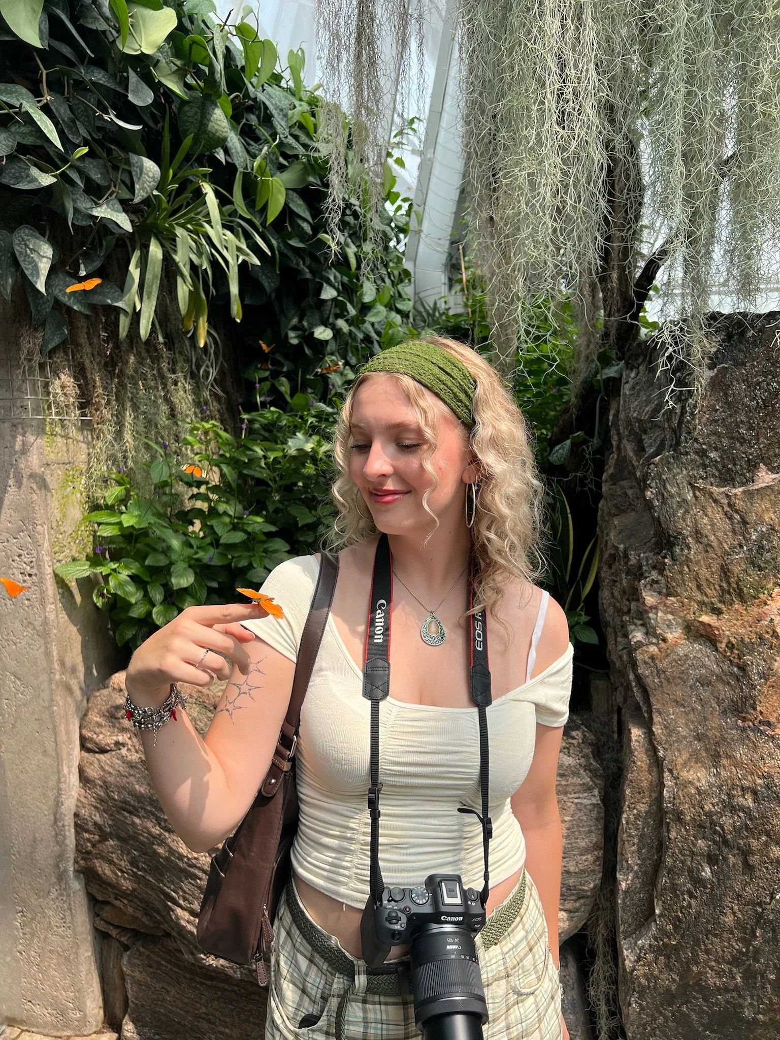 A young woman with blonde curly hair, wearing a green headband, white top, and plaid shorts, is in a lush greenhouse with various green plants and moss-covered rocks, holding a butterfly on her finger and smiling.
