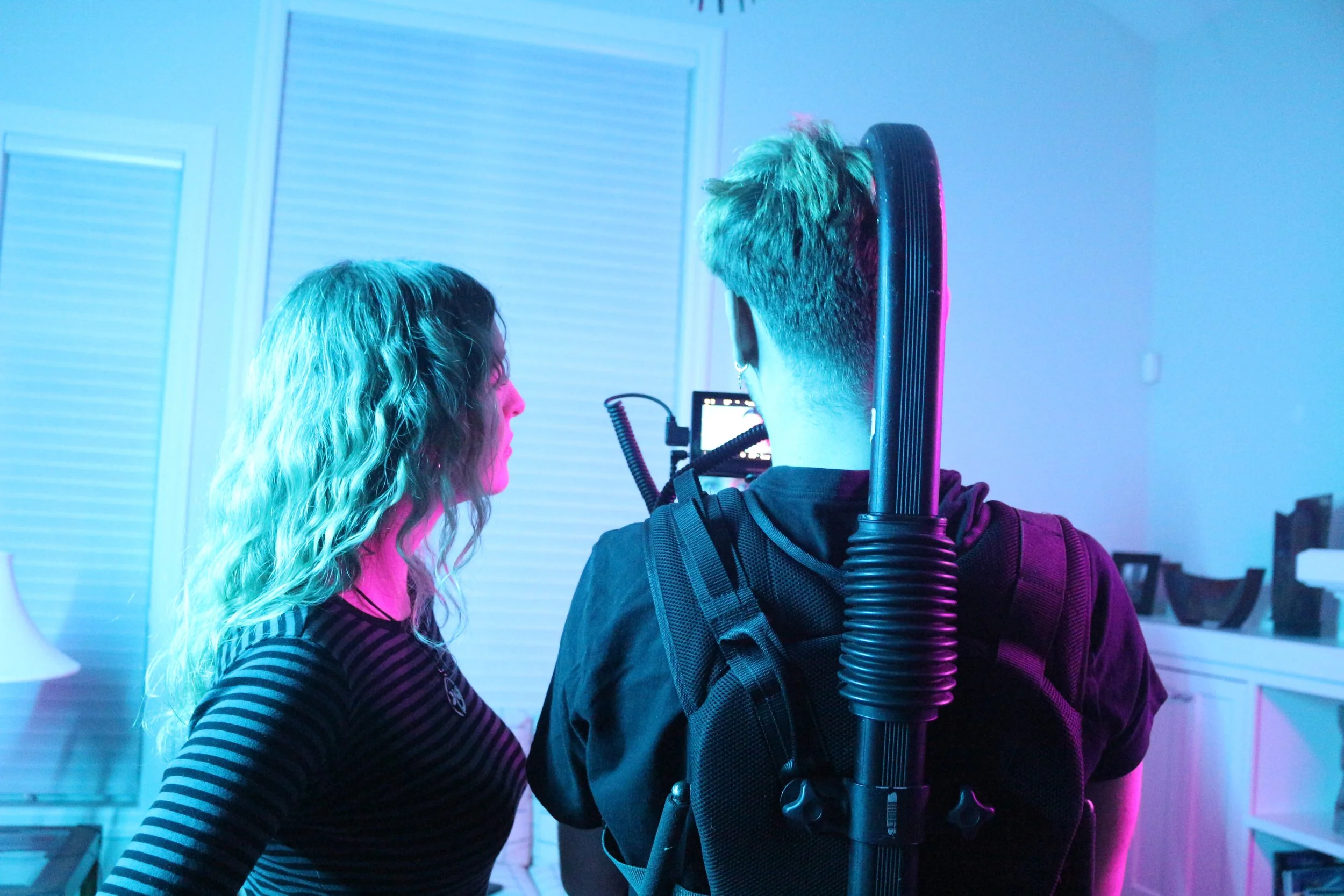 Two people filming a video in a room with blue and pink lighting. One woman with wavy hair and a striped shirt and a man with a backpack and short hair holding a camera.