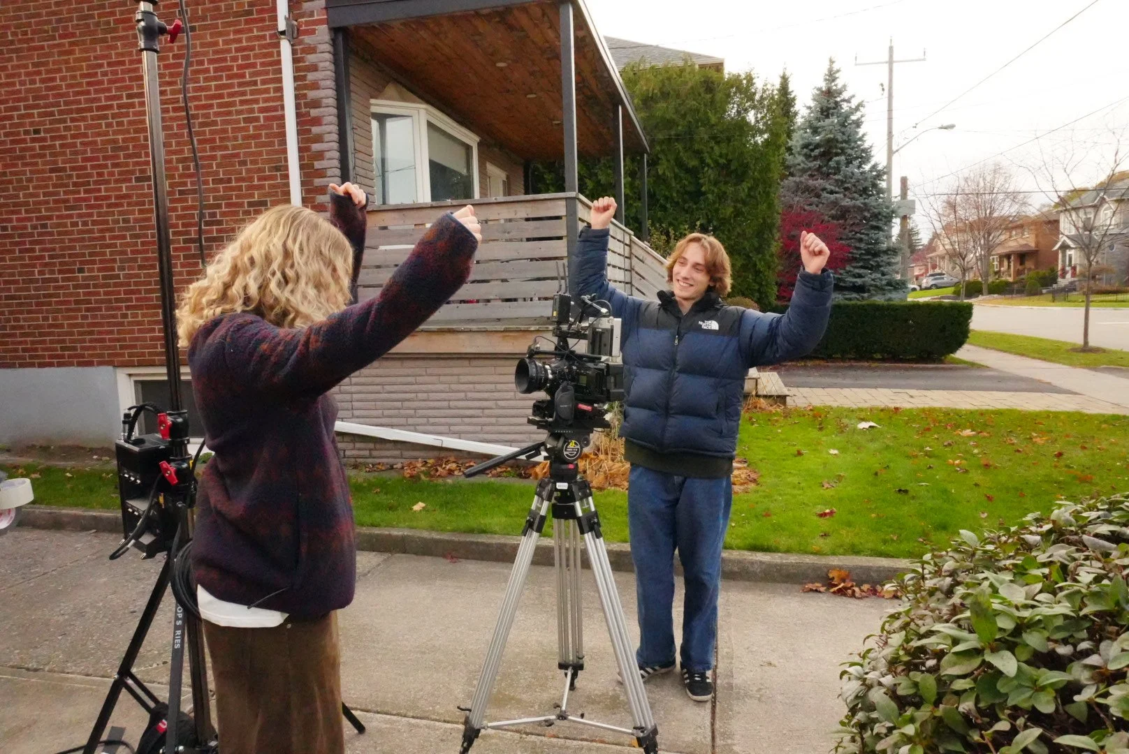 Two young people filming a video outdoors on a cloudy day, with one person standing behind a camera on a tripod and the other person facing them with arms raised in excitement, smiling in front of a brick house and green lawn.