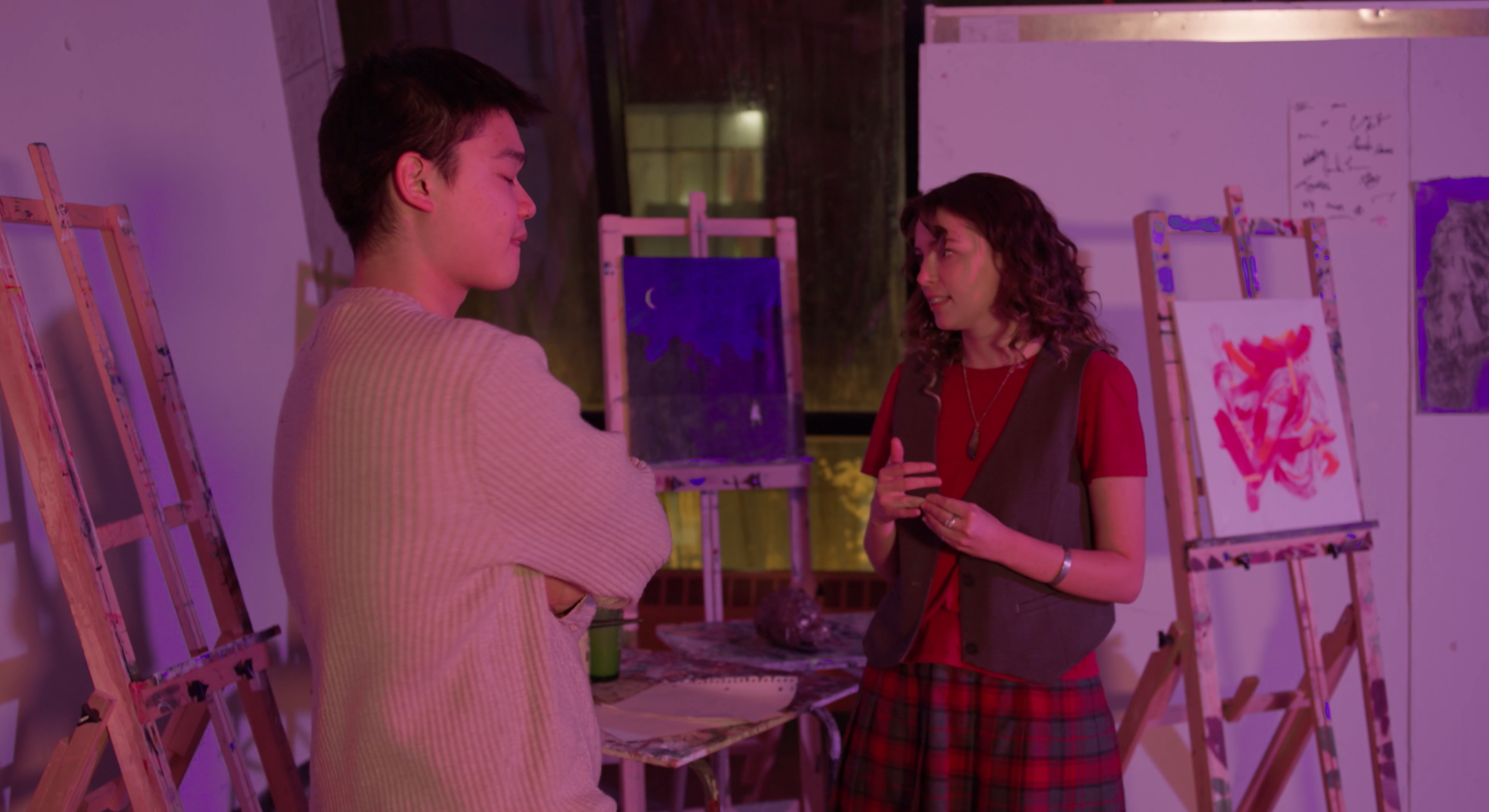 Two women are conversing in an art studio with paintings on easels in the background, illuminated by purple and pink lighting.