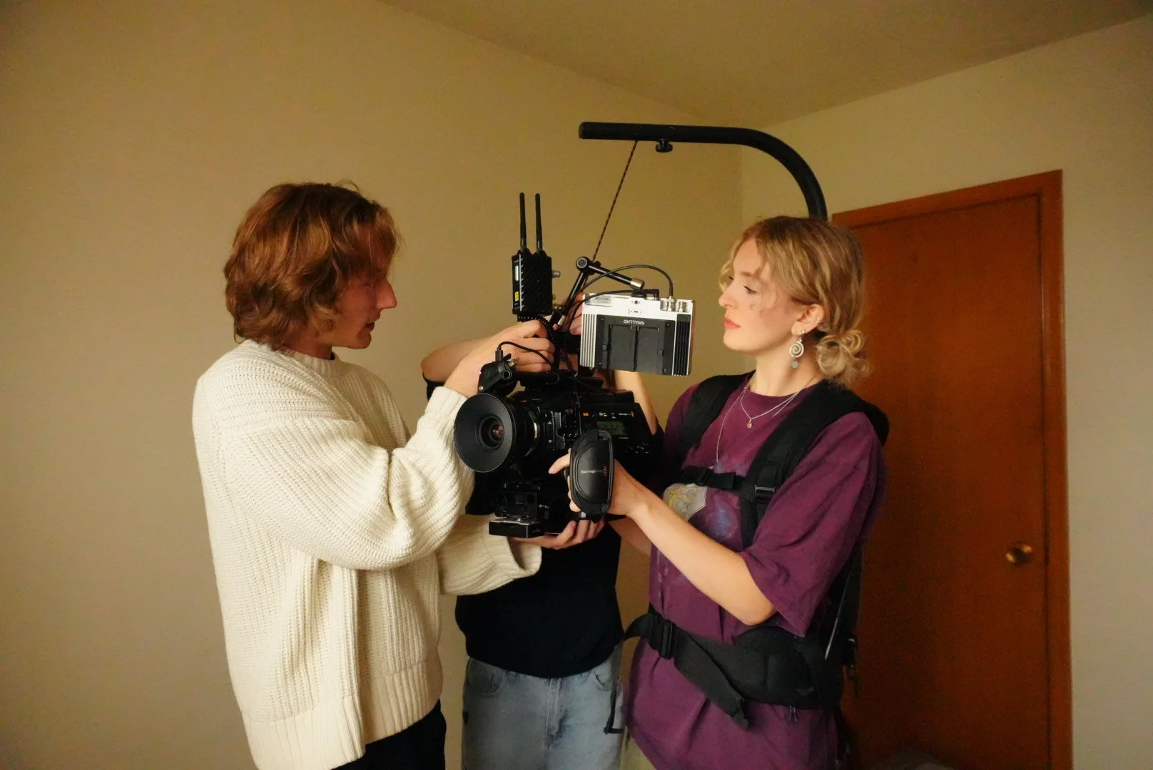 Three people operating a professional film camera in a room with plain walls.