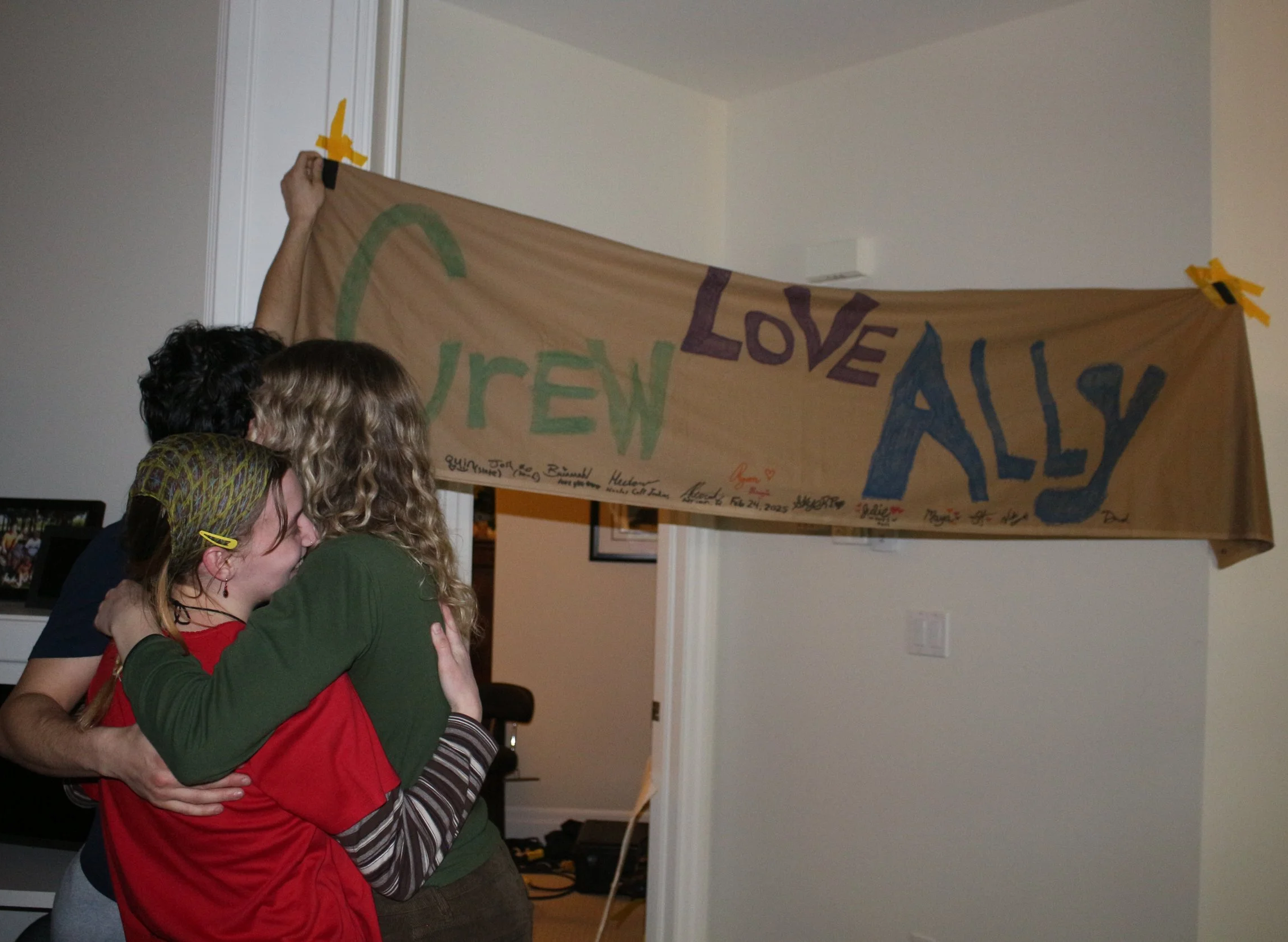 Three people hugging in front of a handmade sign that says "LOVE AILY" with the word "CREW" partially visible, attached to the wall with yellow tape.