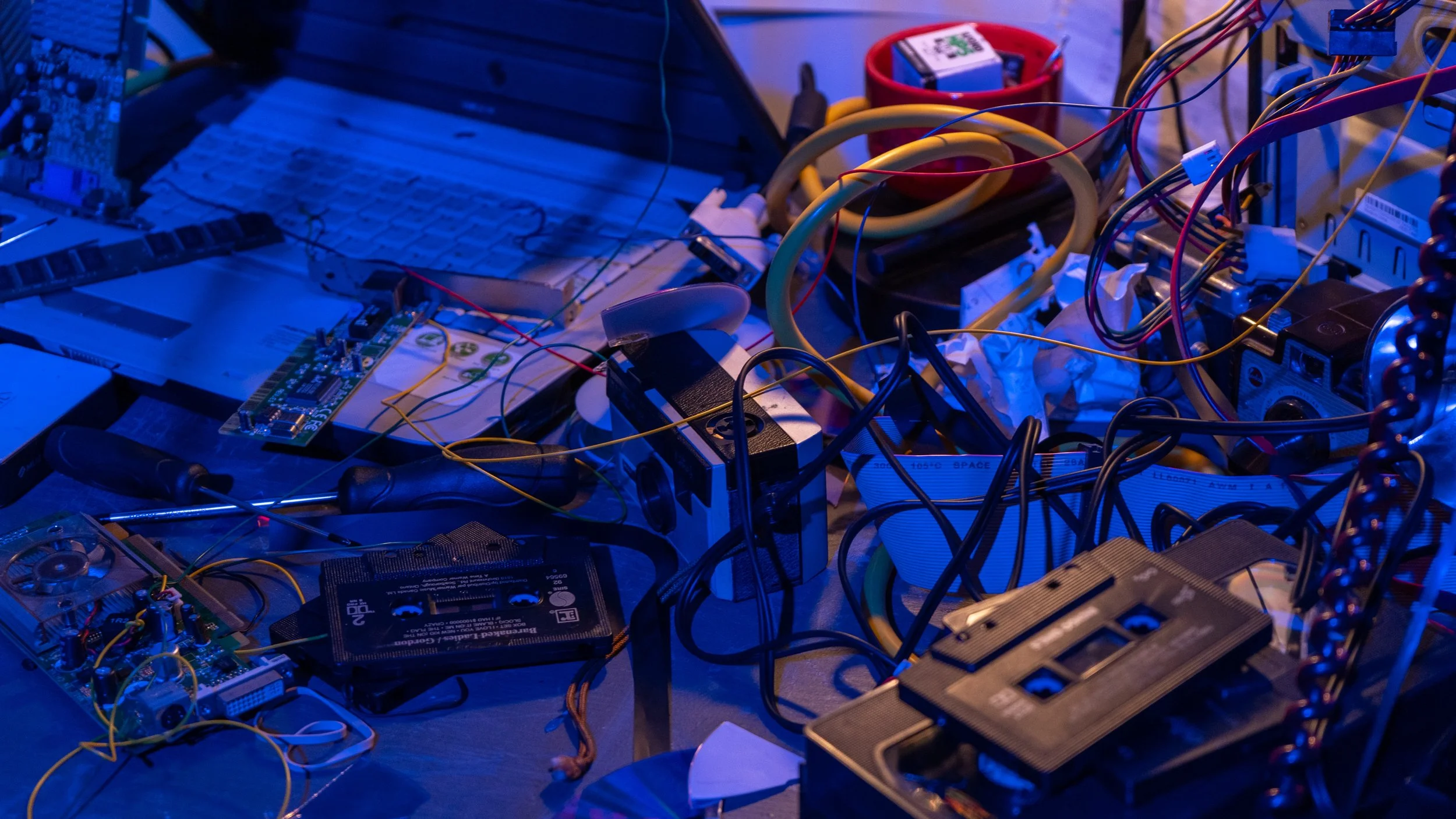 A cluttered workspace with electronic components, wires, tools, and electronic devices, illuminated by blue light.