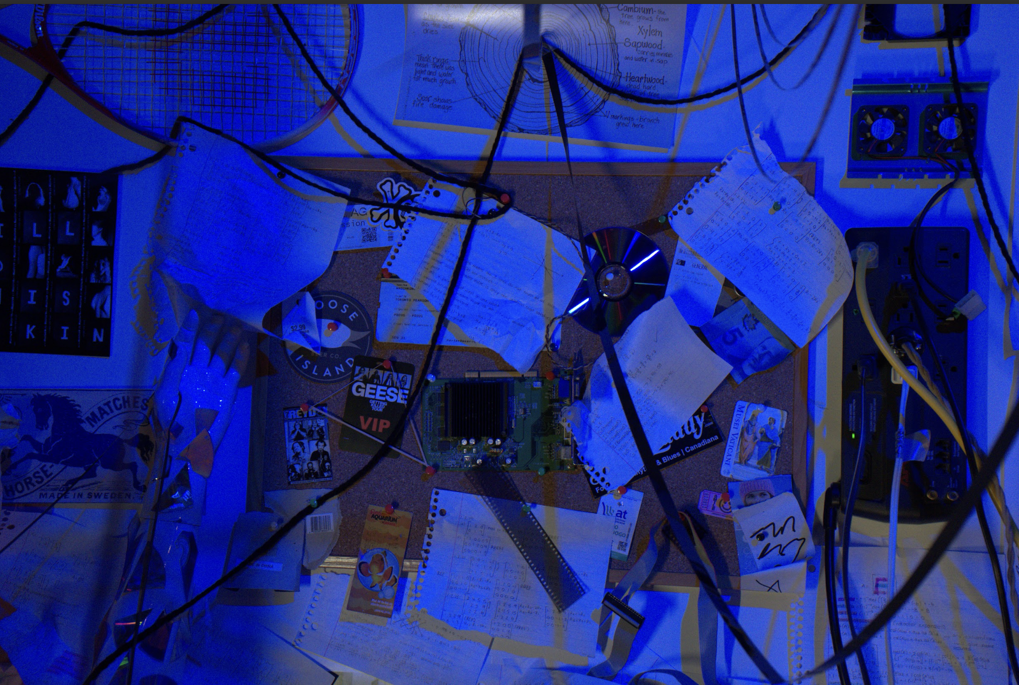A cluttered workspace with scattered papers, electronic components, wires, a circuit board, a CD, and various stationery items illuminated by blue light.