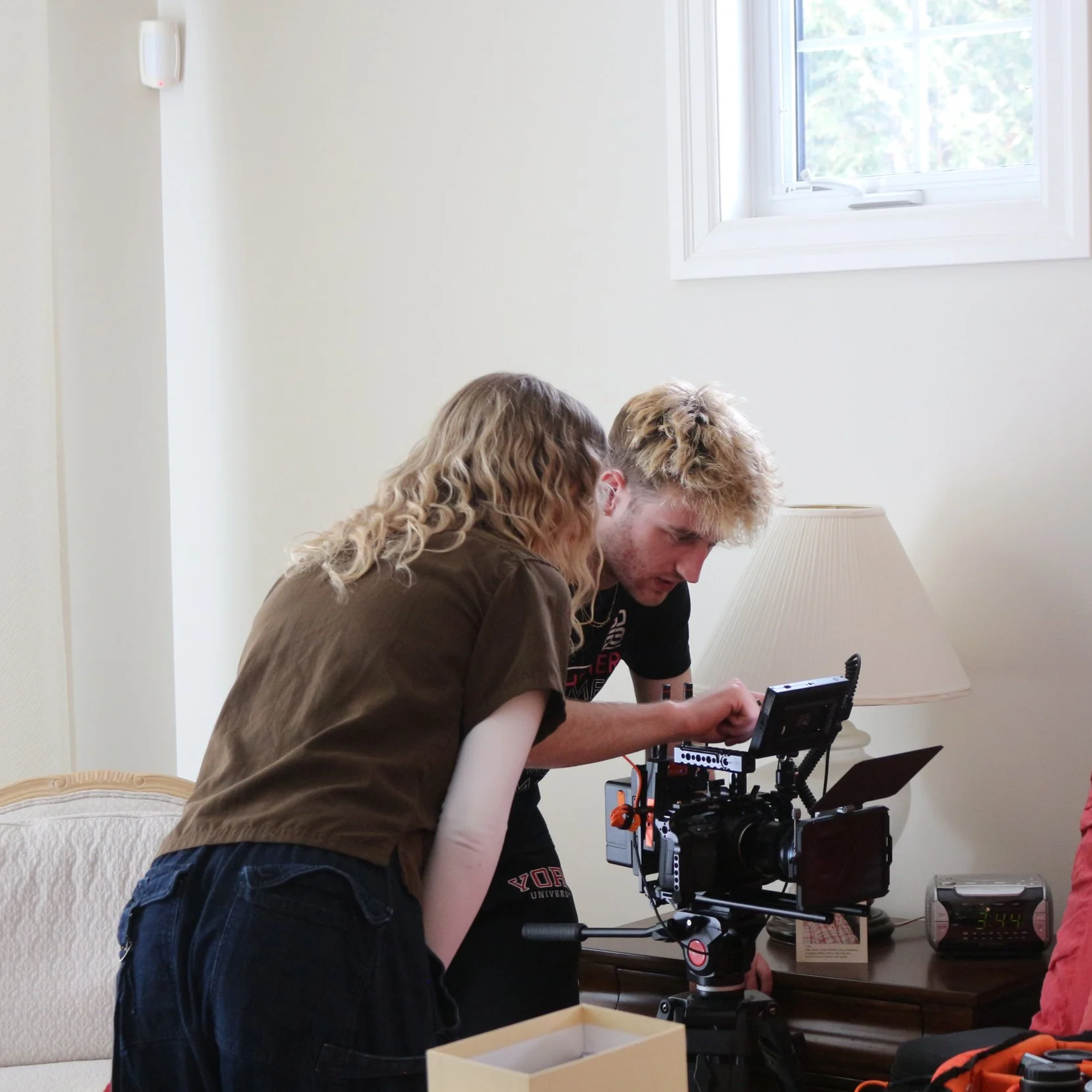 Two people working on a film camera setup in a room with a window and a table lamp.