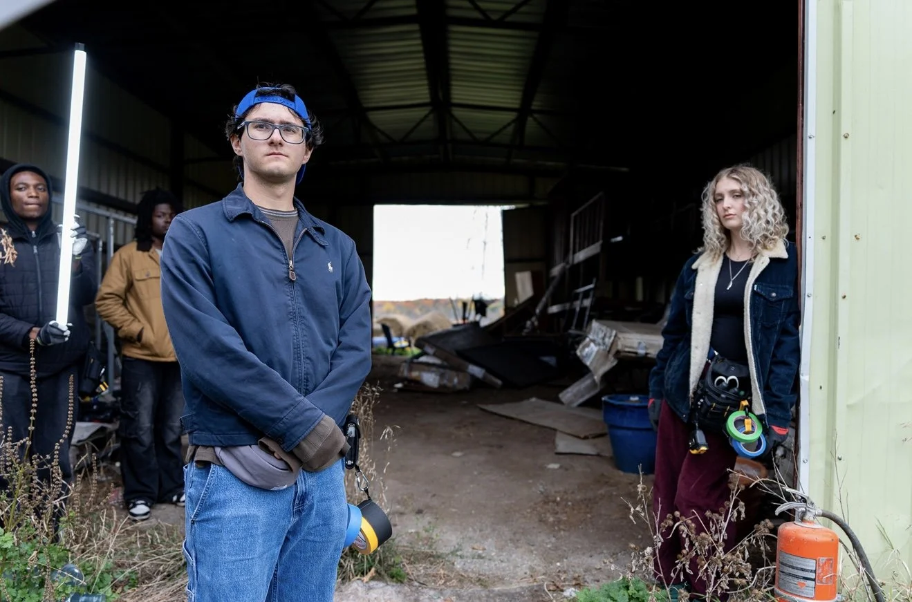 Group of young adults standing inside a partially open shed or garage, with some debris and tools behind them. The man in the foreground wears glasses, a blue cap backwards, and a navy jacket, holding a measuring tape. The woman on the right has curly blonde hair, a black shirt, a denim jacket with a shearling collar, and a tool belt with colorful rings, and stands near a fire extinguisher. Two other individuals are in the background.