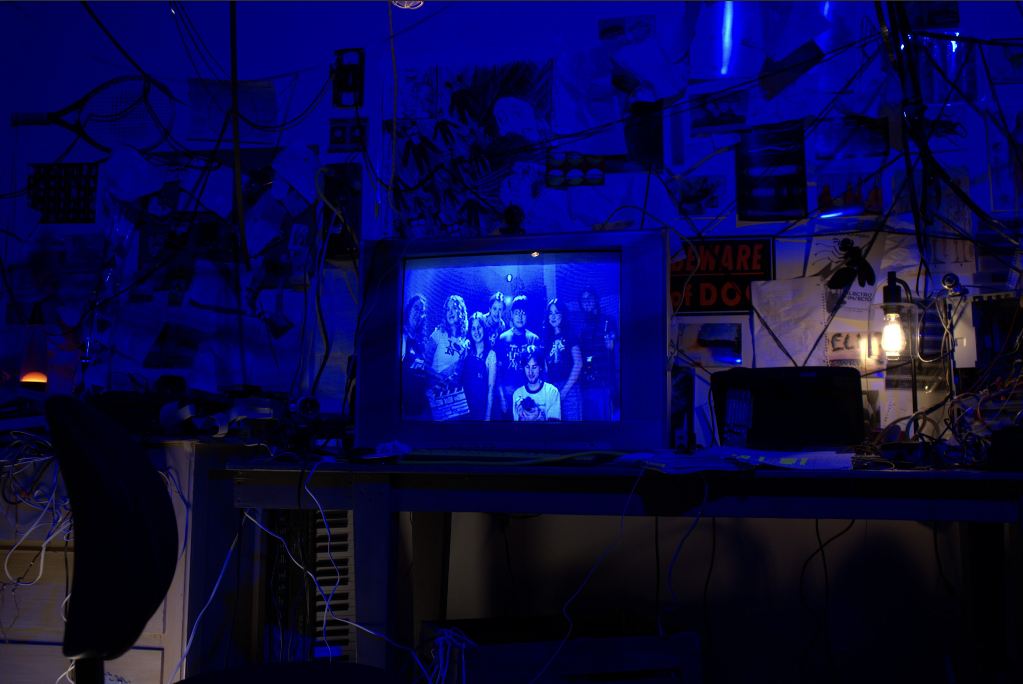 A cluttered workspace with a computer monitor displaying a group photo, surrounded by tangled wires, posters, and a lit lantern on the right.
