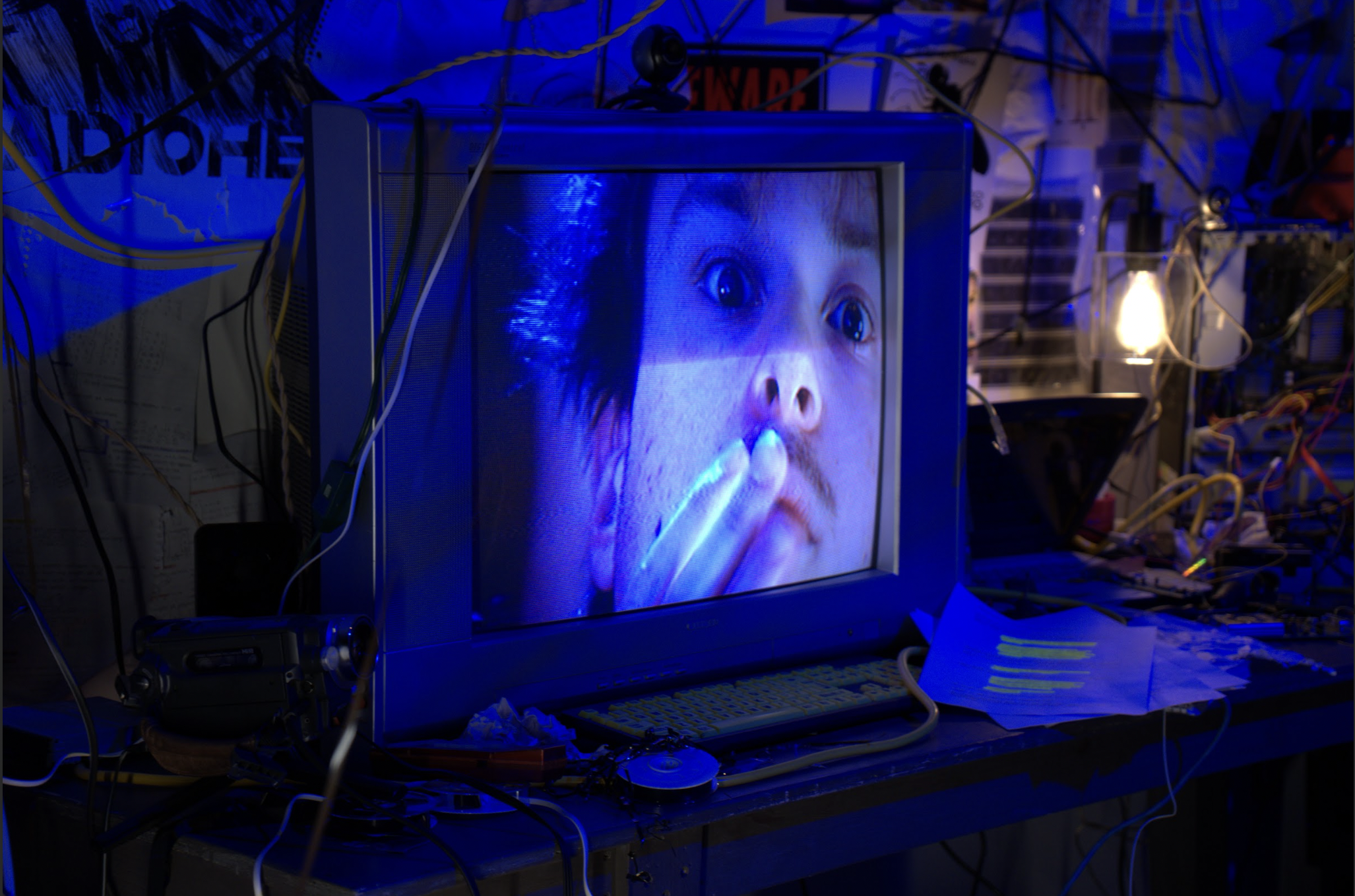 An old television monitor displaying the face of a person with wide eyes and a hand near their mouth. The surrounding area shows an cluttered workspace with wires, papers, and a lamp, lit by a small light bulb.
