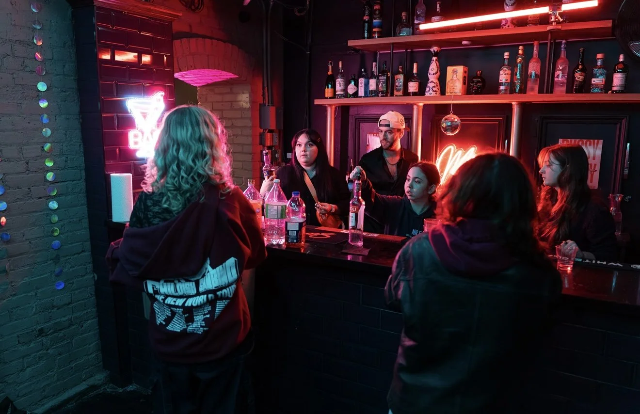 A group of six young people gathered at a bar counter with colorful neon lighting, drinking and socializing in a dimly lit venue.