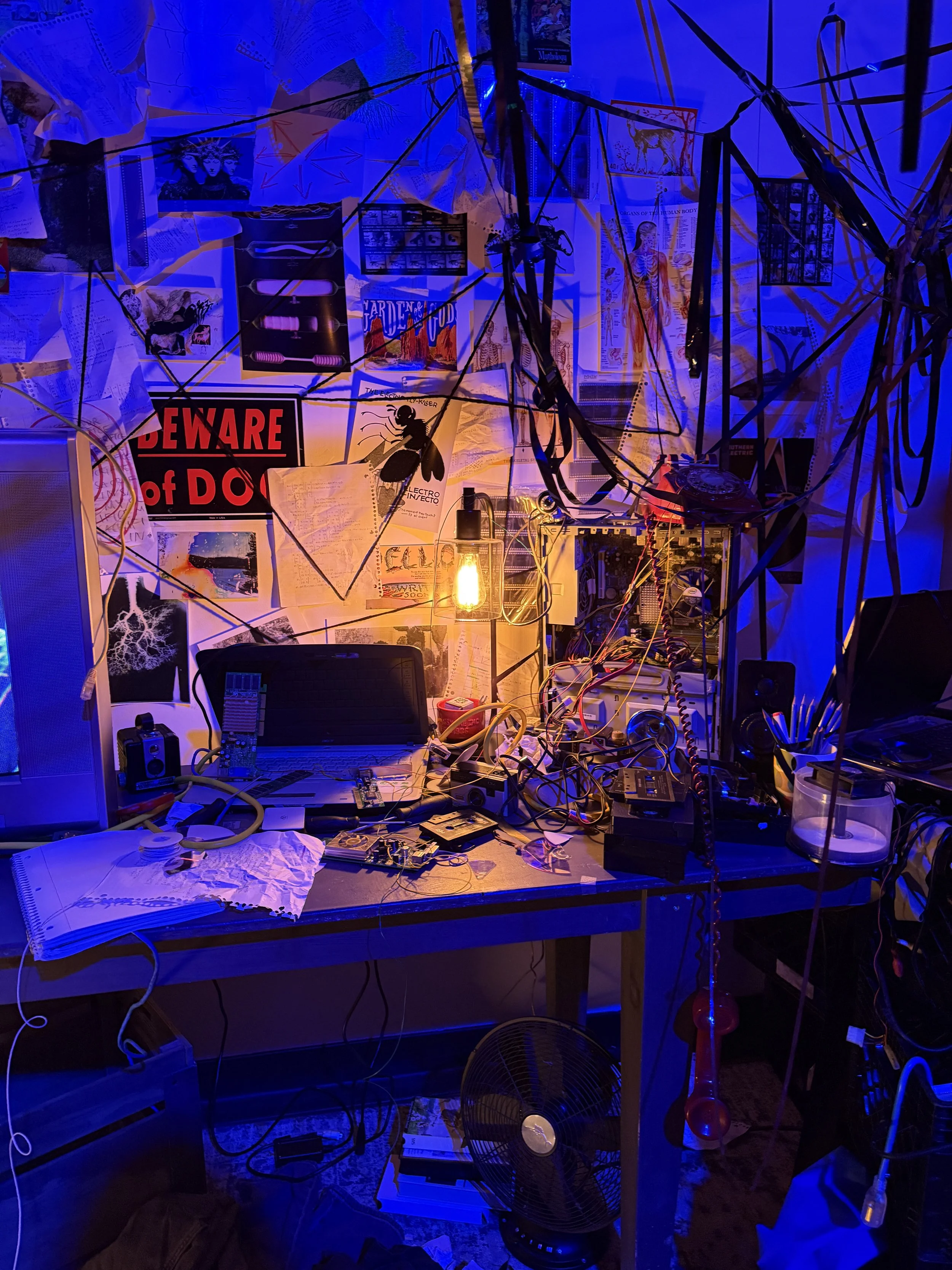 A cluttered computer workspace with wires, papers, and electronic components, illuminated by a yellow Edison bulb and surrounded by posters and notes on the wall.