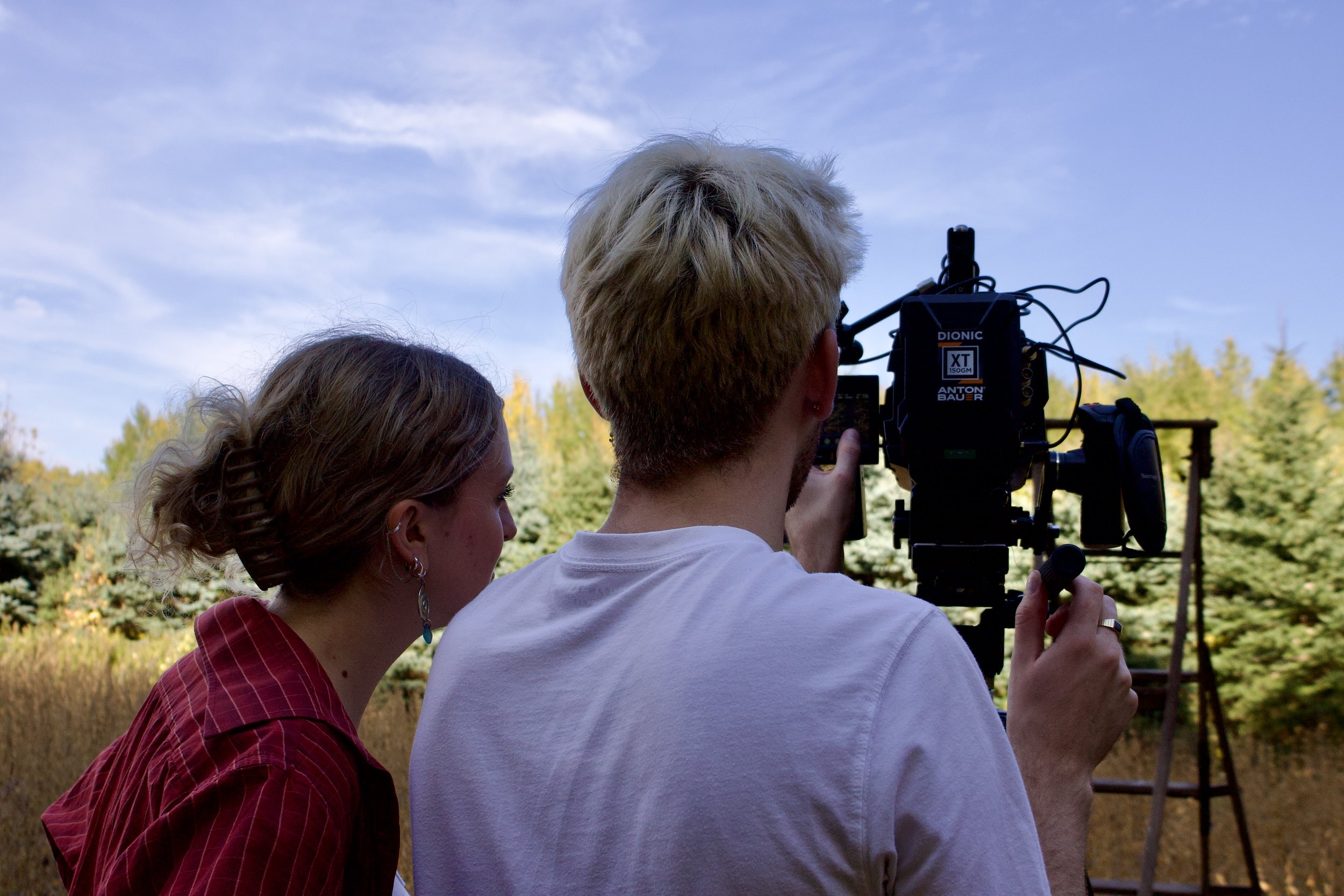 Two people filming outdoors with a camera, greenery and a blue sky in the background.