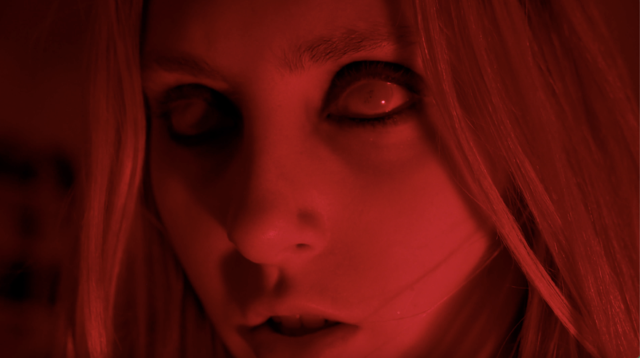 Close-up of a person with long red hair illuminated by red light, with intense eyes and a serious expression.