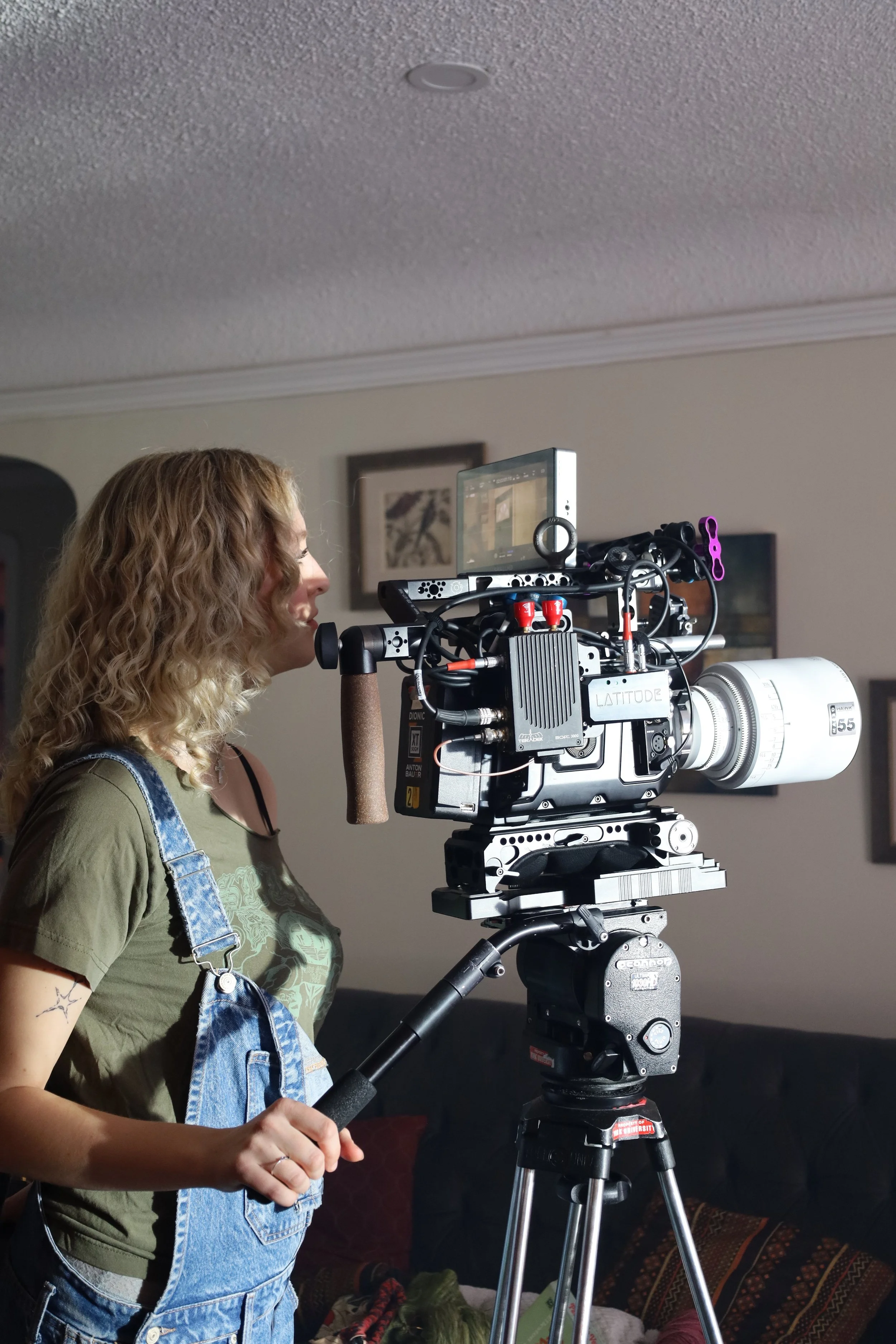A woman with curly blonde hair operating a professional film camera on a tripod indoors.