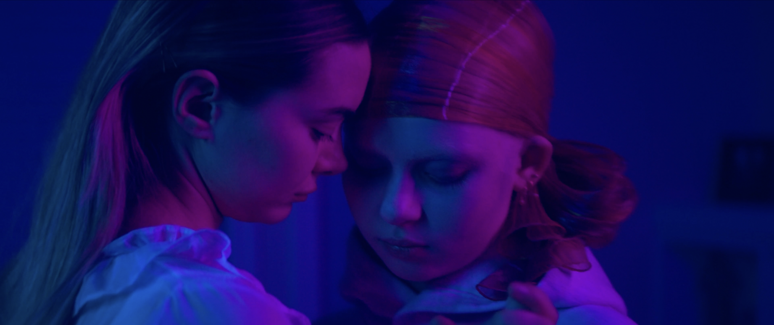 Two women with closed eyes and solemn expressions, closely embracing in a dimly lit room with neon purple and blue lighting.