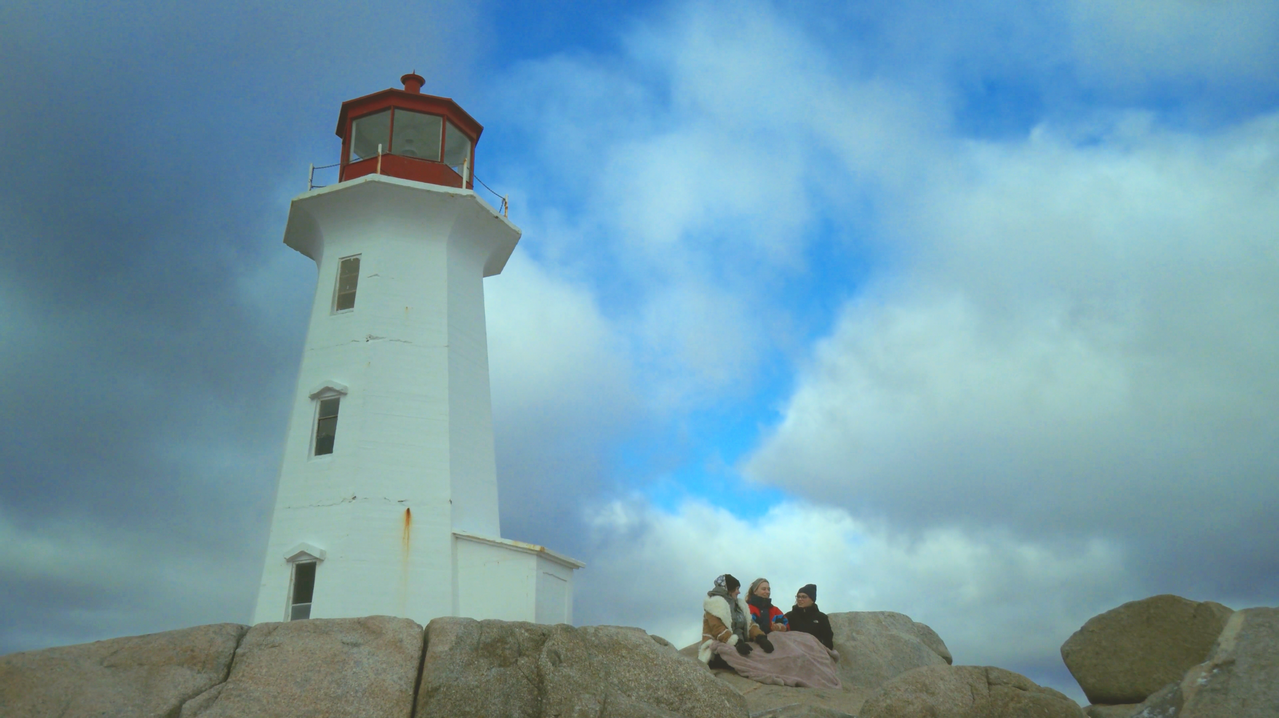 A white lighthouse with a red top stands against a partly cloudy sky. Three people sit on rocks near the lighthouse, bundled in warm clothing.