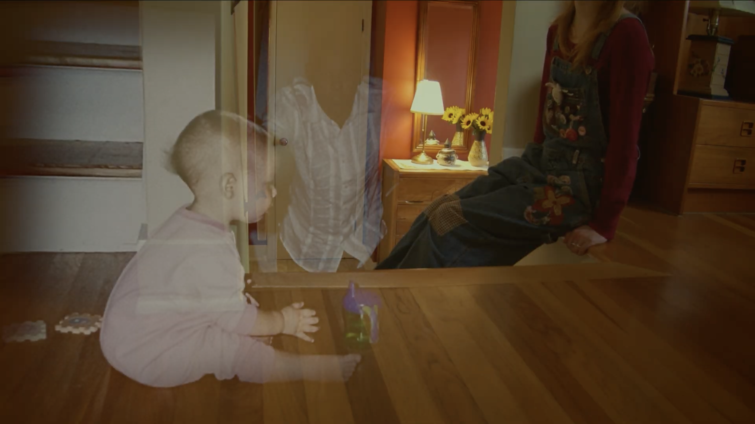 Two children and a woman are separated by a glass door. The girl is sitting on the floor, and the boy is on the other side of the glass, standing. The woman is sitting on the edge of a table, looking at the girl.