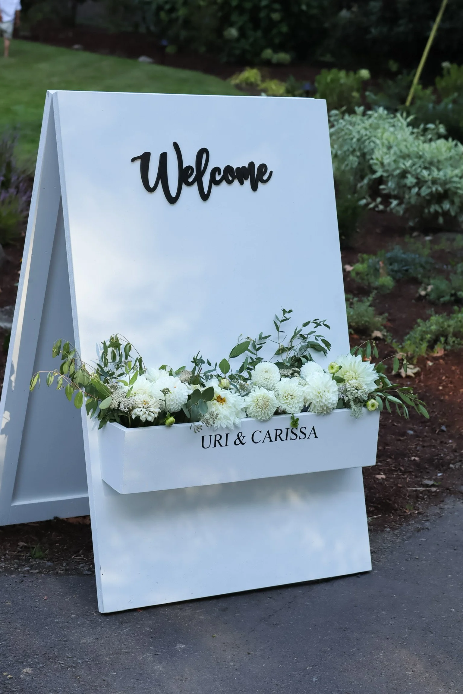 White wedding sign with the word 'Welcome' at the top, decorated with fresh white flowers and greenery, with the names 'Uri & Carissa' written on a flower box below. The sign is outdoors on a paved area with a garden in the background.