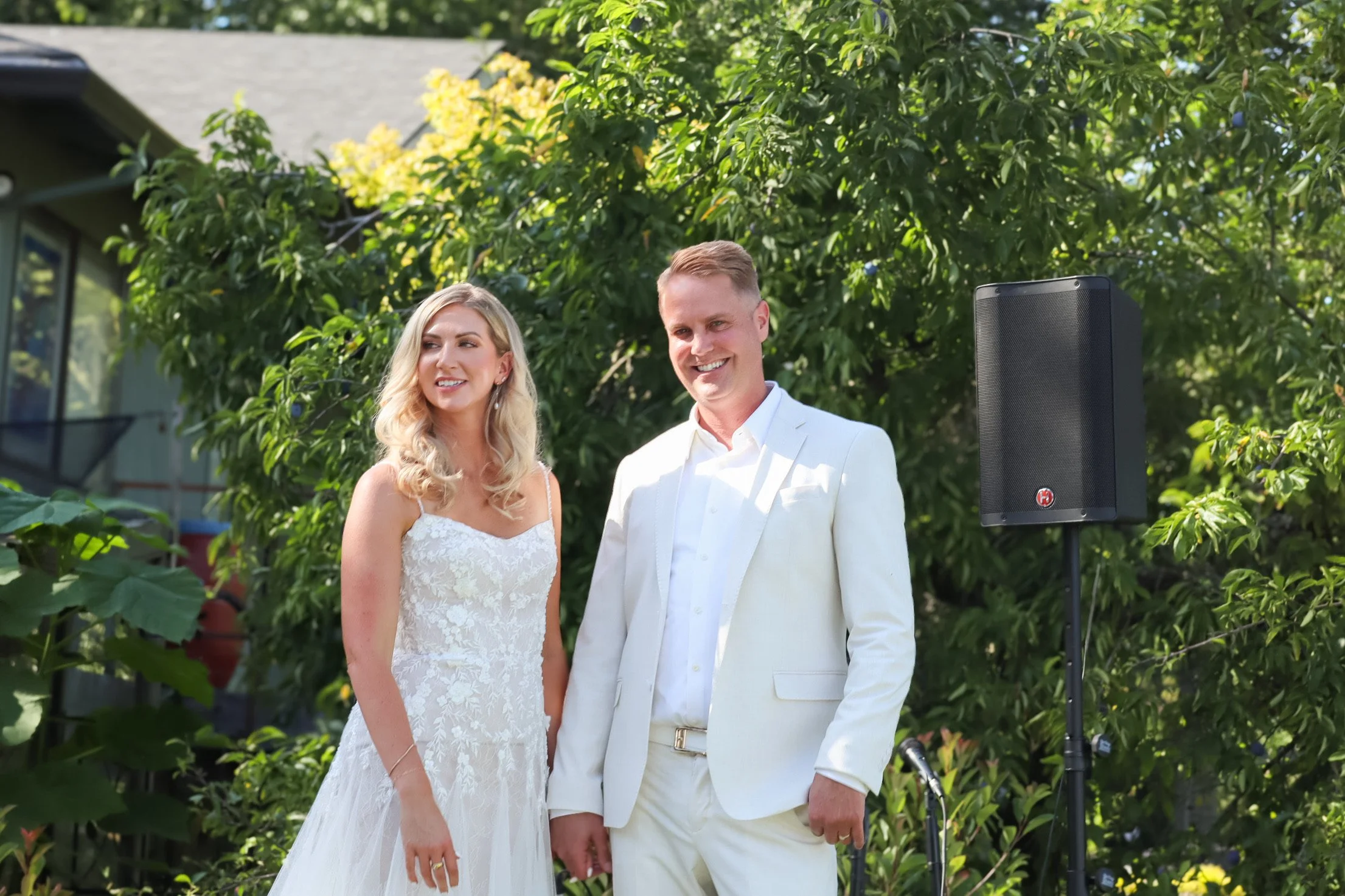 A smiling bride in a white lace wedding dress standing outdoors next to a groom in a white suit, with green trees in the background and a black speaker on a stand nearby.