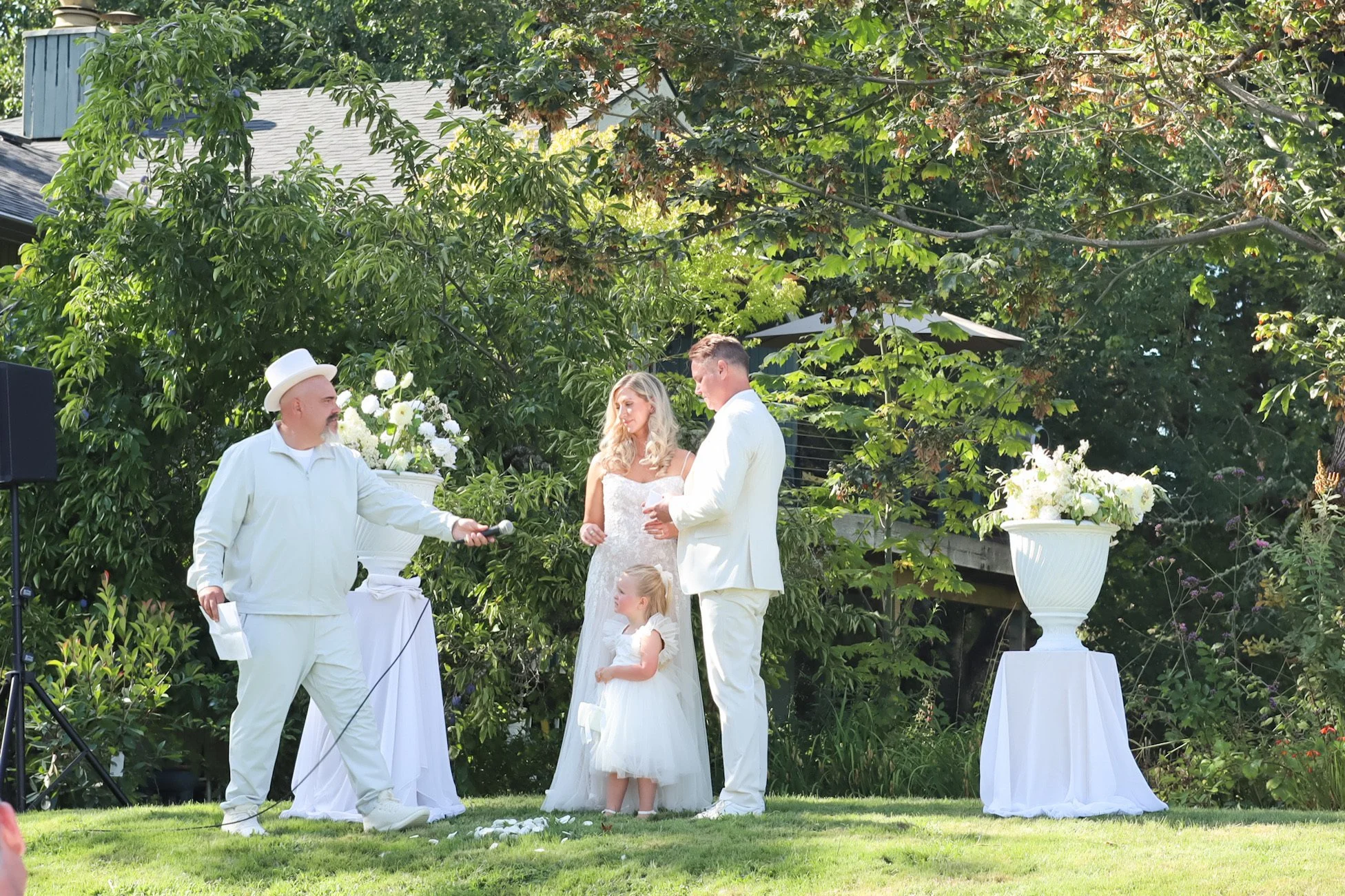 A wedding ceremony taking place outdoors with a bride and groom standing in front of an officiant. The bride is wearing a white lace dress, and the groom is in a white suit. There is a young girl in a white dress standing between them. The officiant 
