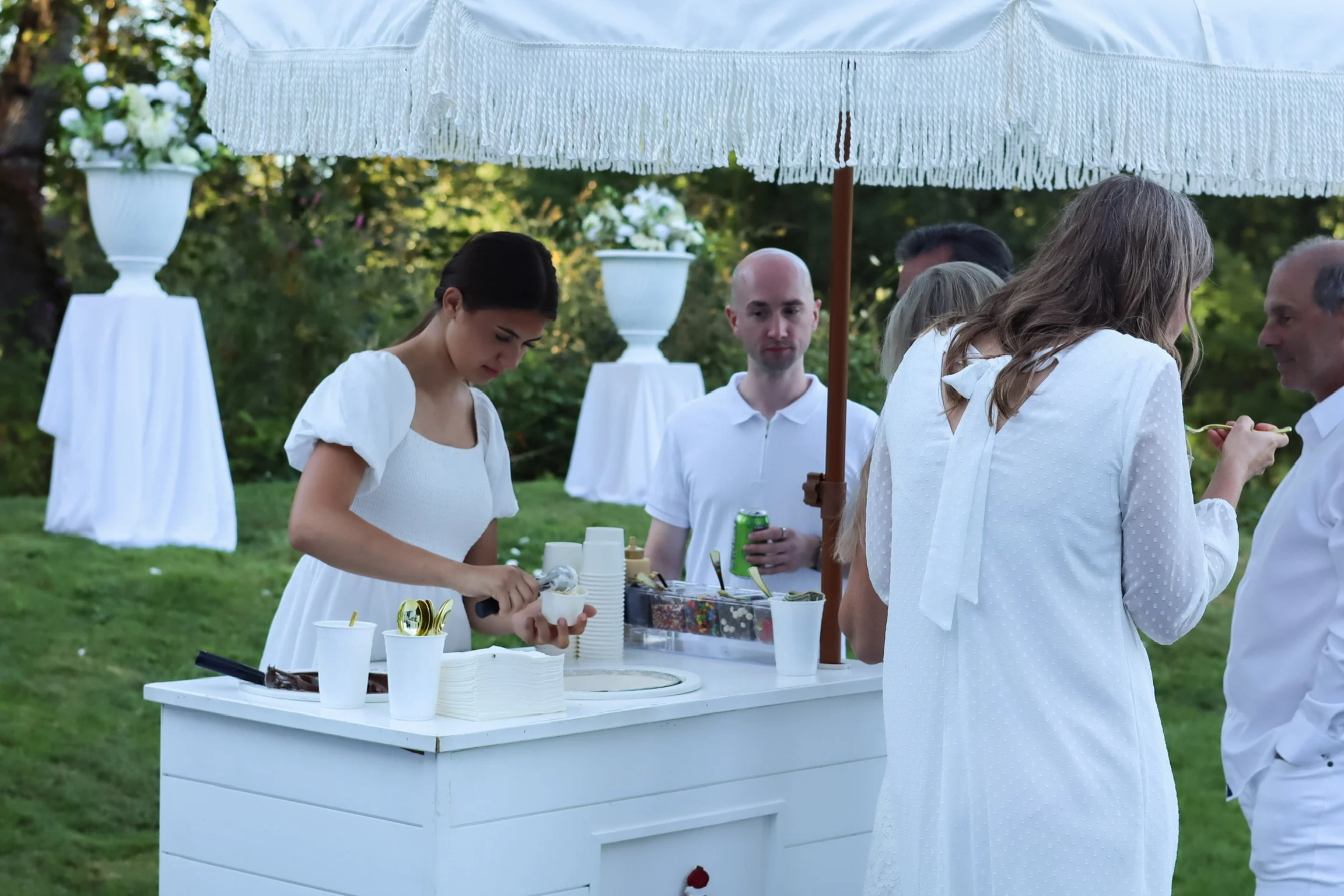 People gathered under a large white umbrella at an outdoor event, with a woman serving ice cream from a white cart, and others socializing in the background.